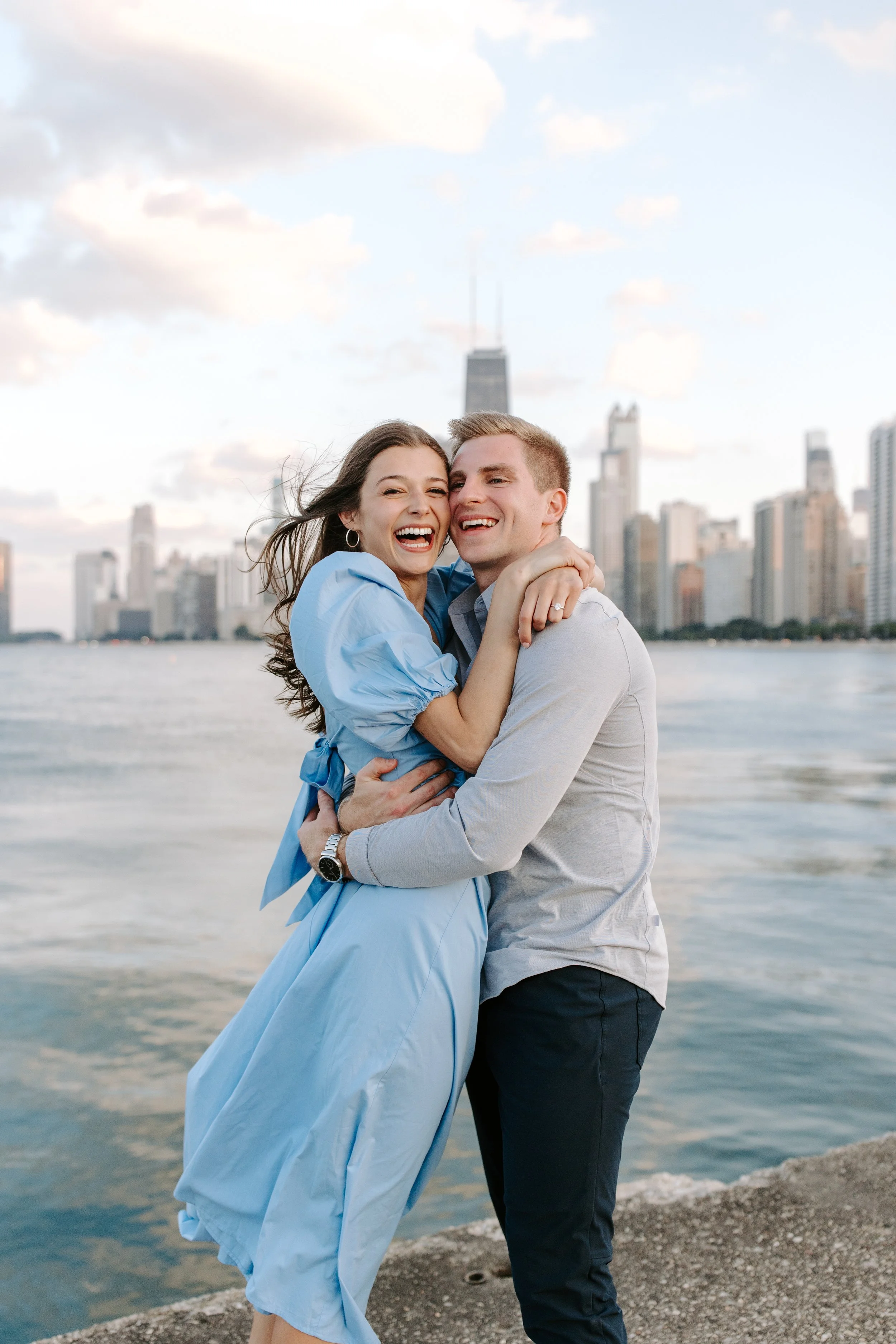 A happy couple embracing each other near a body of water with a city skyline, including a tall building with antennas, in the background during sunset or late afternoon.