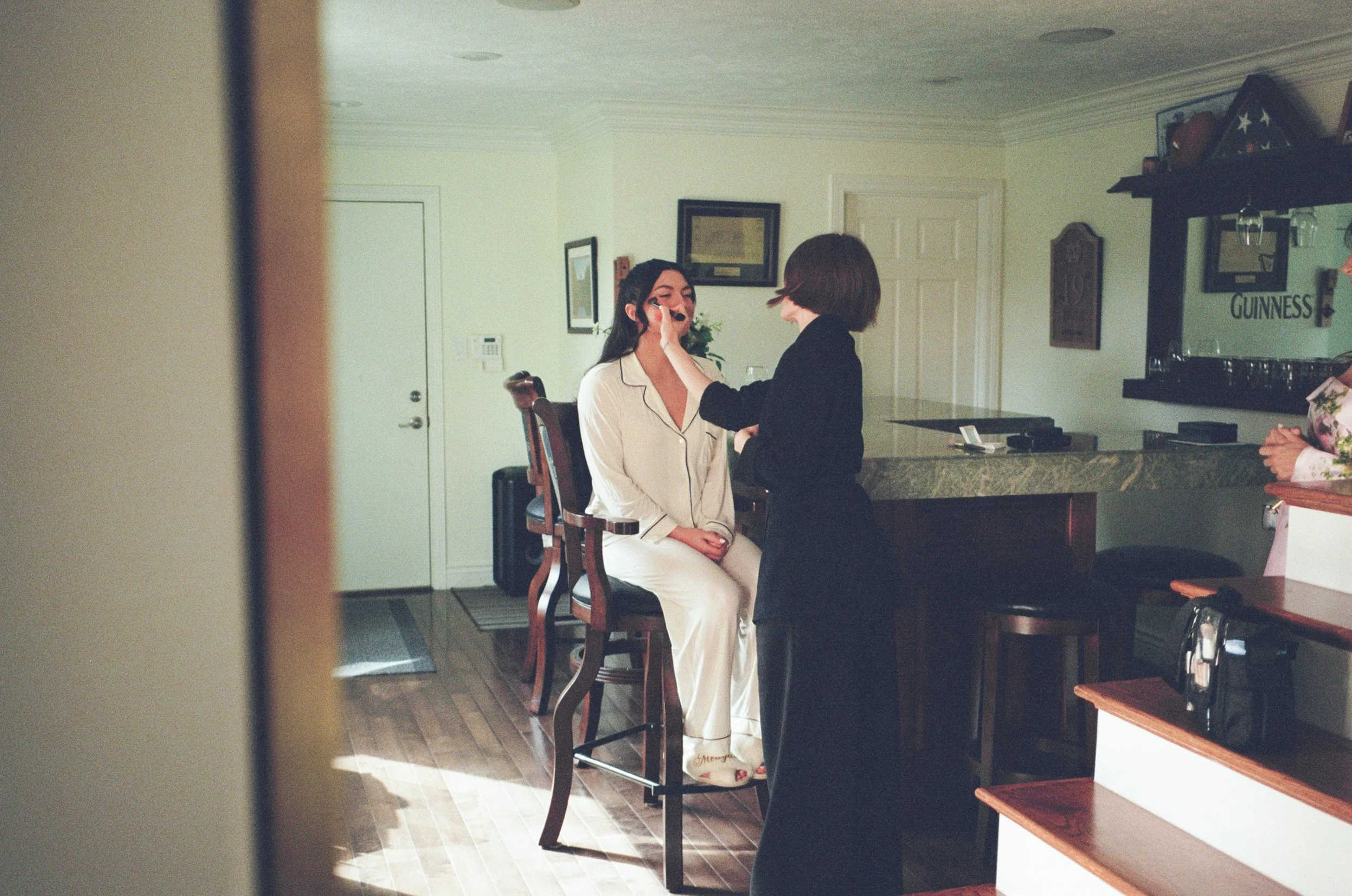 A woman in pajamas sits on a barstool while another woman applies makeup to her face in a room with a bar and framed pictures on the wall.