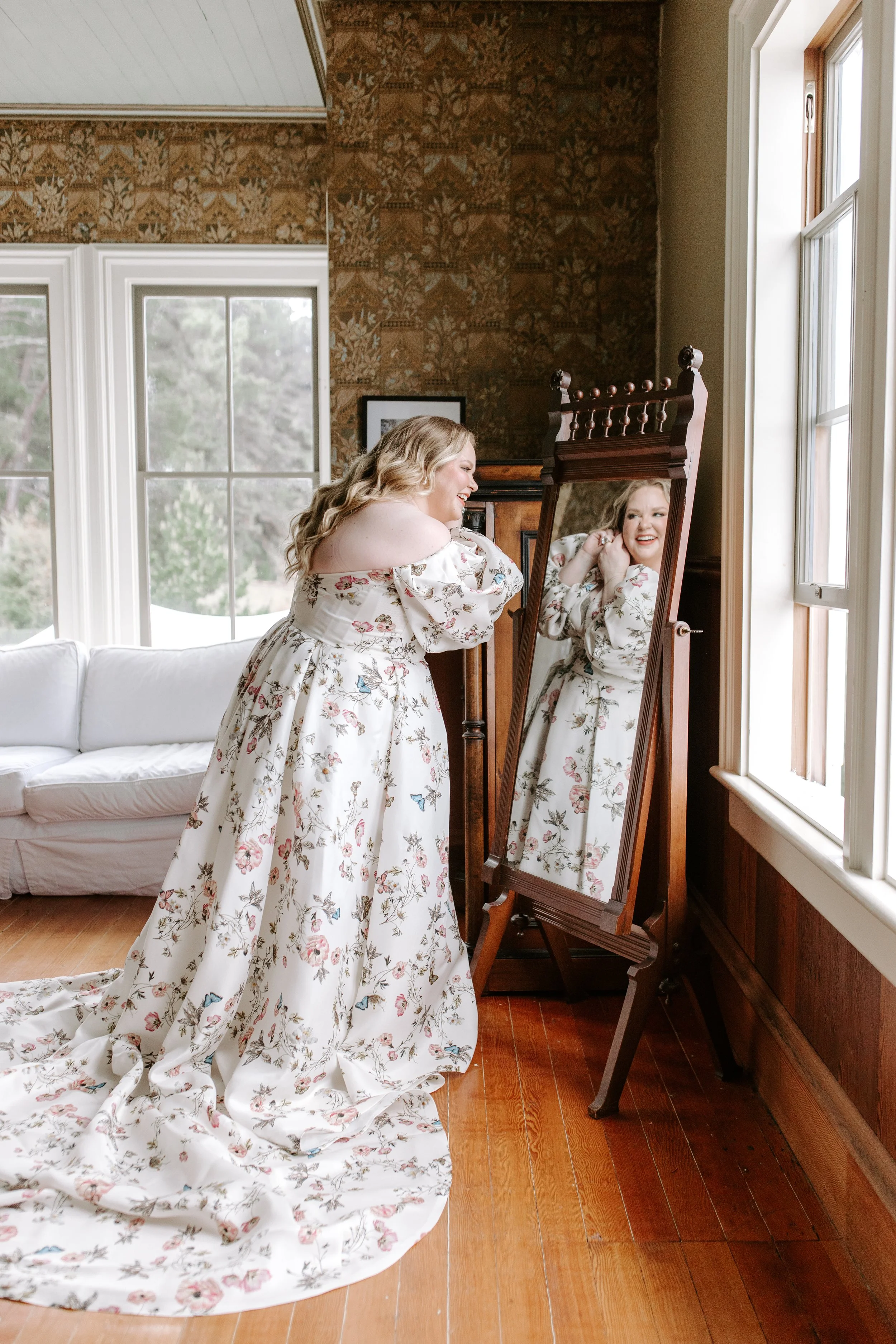 A woman with long blonde hair wearing a white floral wedding dress is looking at herself in a tall wooden mirror, smiling and adjusting her earrings inside a room with large windows, a white couch, and a wooden floor.