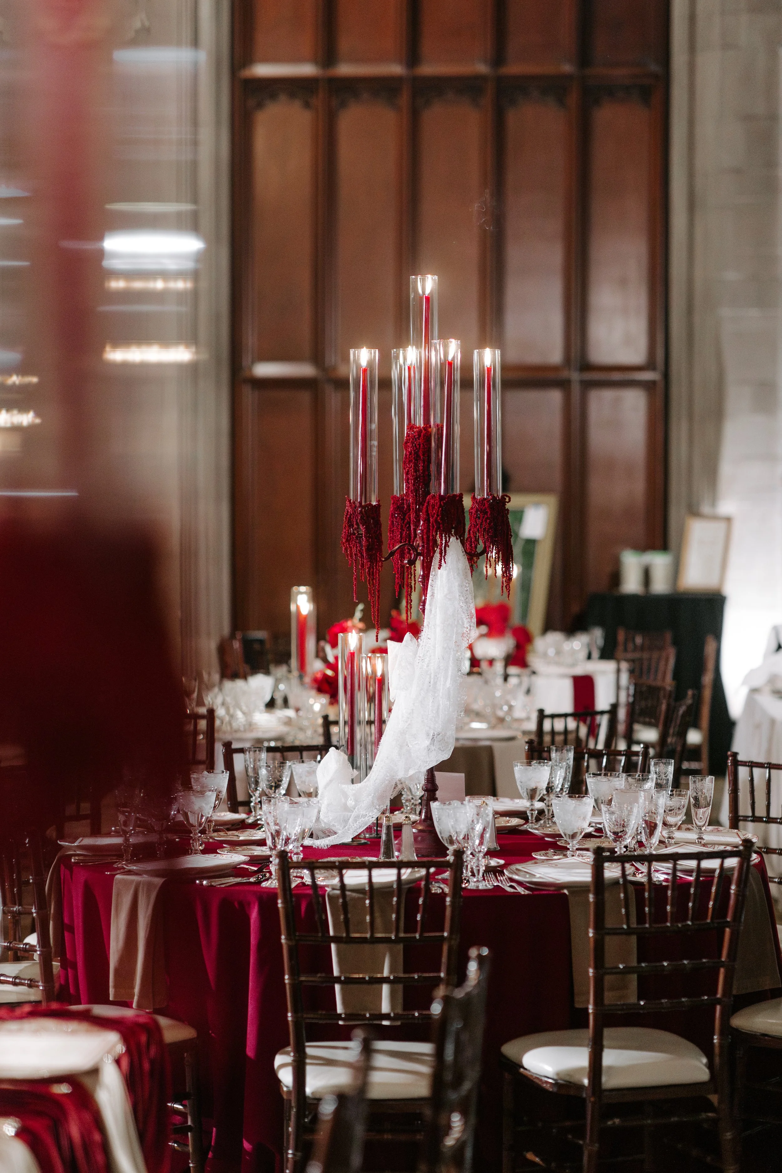 A banquet table set for a formal event, featuring a tall centerpiece with multiple glass candle holders and red decorative elements, white tablecloths, glassware, and chairs with dark wood frames and white cushions, in an elegant room with wooden wal