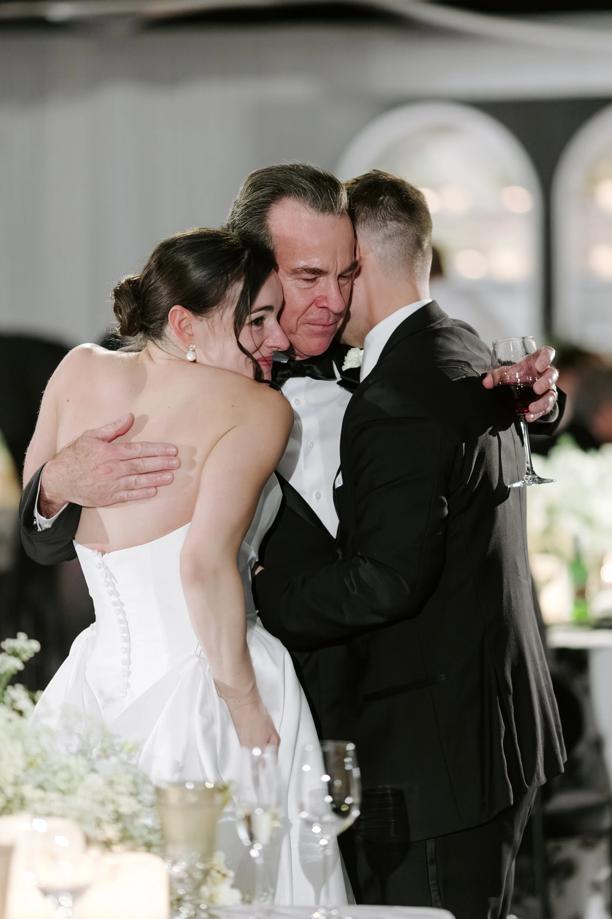 A group of three people, a man and two women, hug each other at a wedding reception. The bride, in a white wedding dress, is embracing the older man, who is in a tuxedo, and the groom, in a black tuxedo, is holding a glass of red wine. They are insid