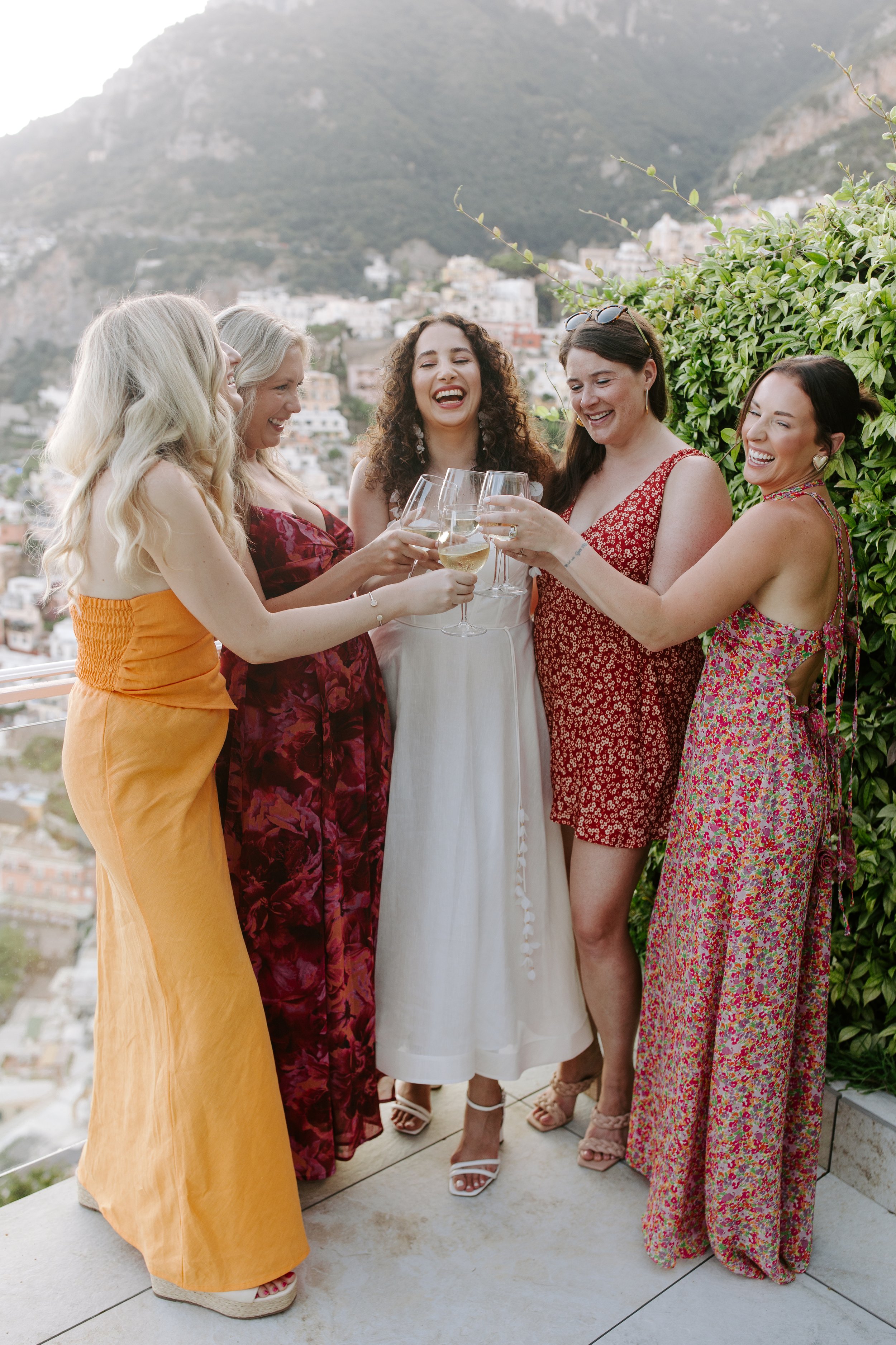 A group of five women gathered outdoors on a balcony, smiling and holding glasses of white wine. They are dressed in colorful, summery dresses. The background features a hilly landscape with buildings and greenery.