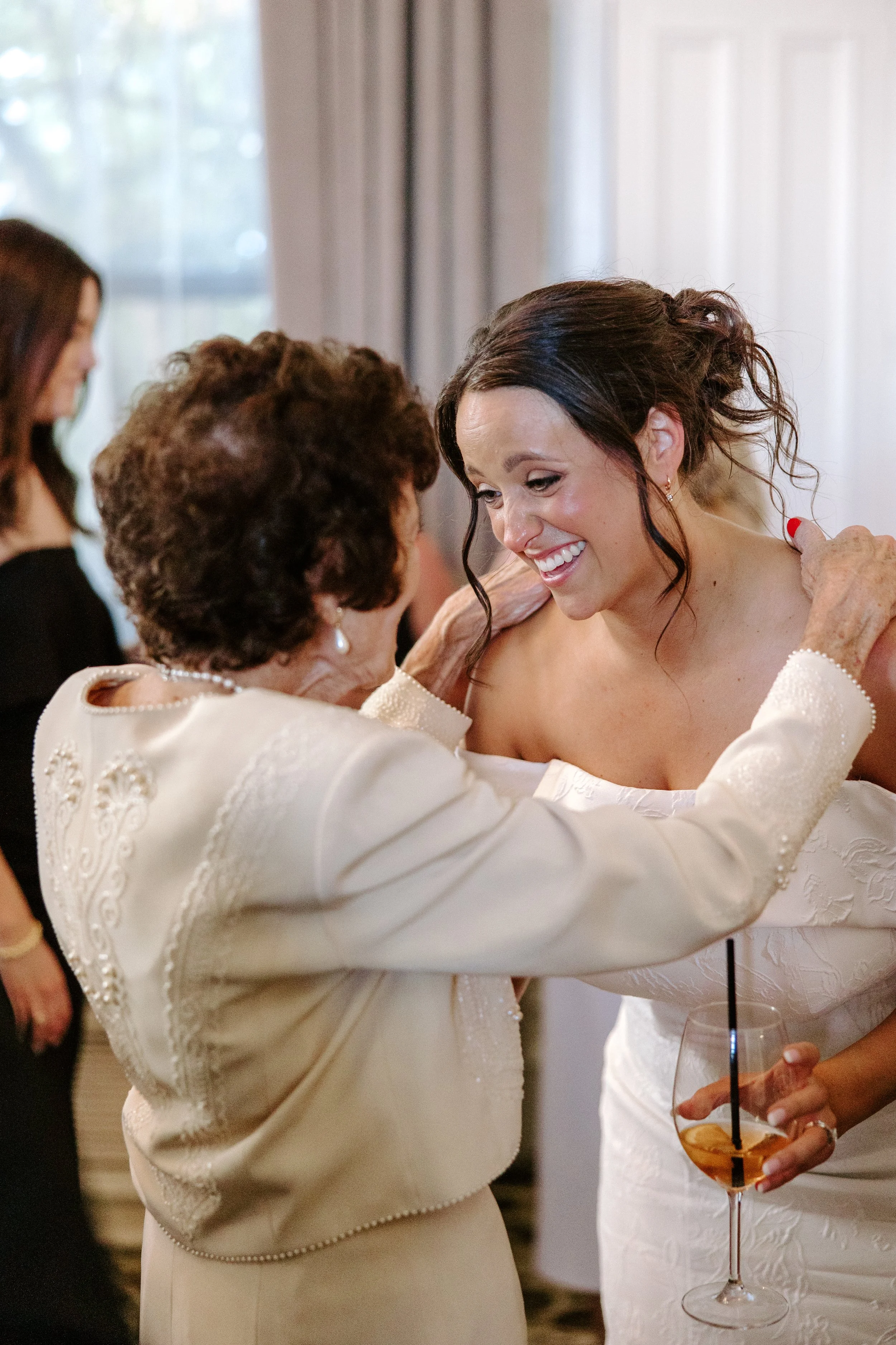 Two women, one older and one younger, are sharing a warm hug at a social gathering, with the younger woman holding a glass of wine with an ice cube and straw. The women are dressed in elegant, light-colored clothing, and there are other people in the