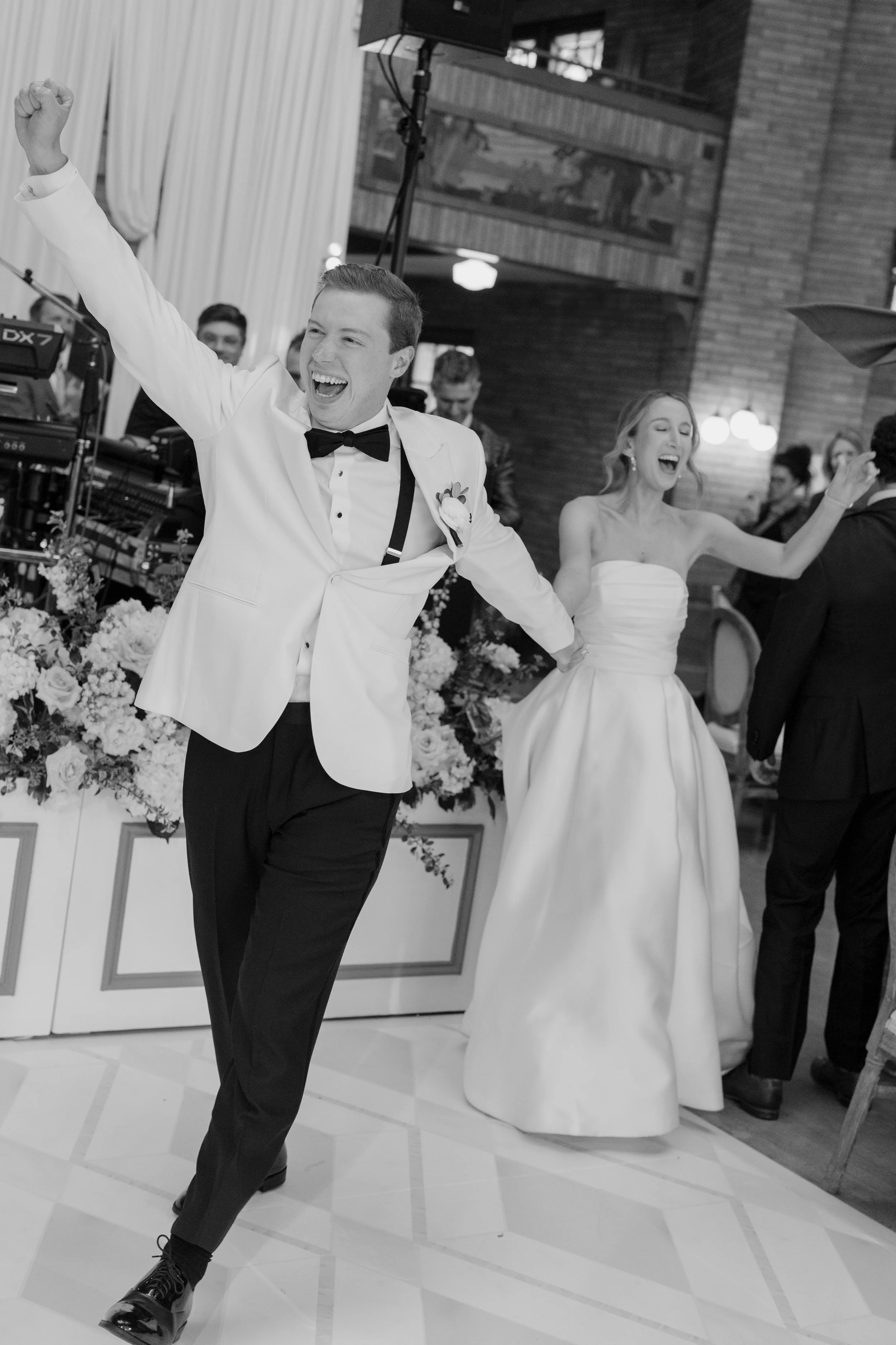 A man and woman dressed in wedding attire dancing at a wedding reception, smiling with their arms raised.