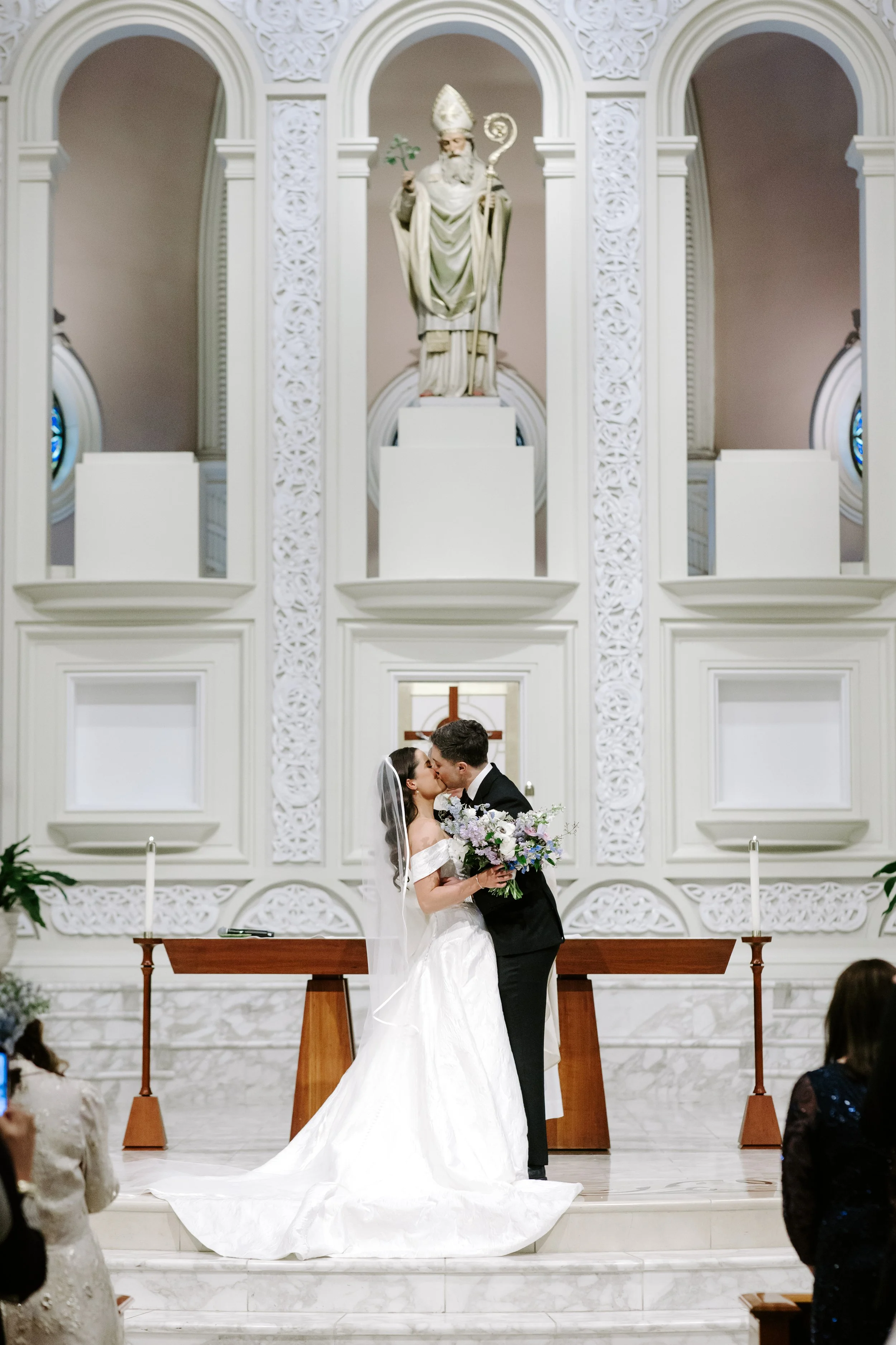 A bride and groom share a kiss during their wedding ceremony inside a church with white ornate walls and a large religious statue above them.