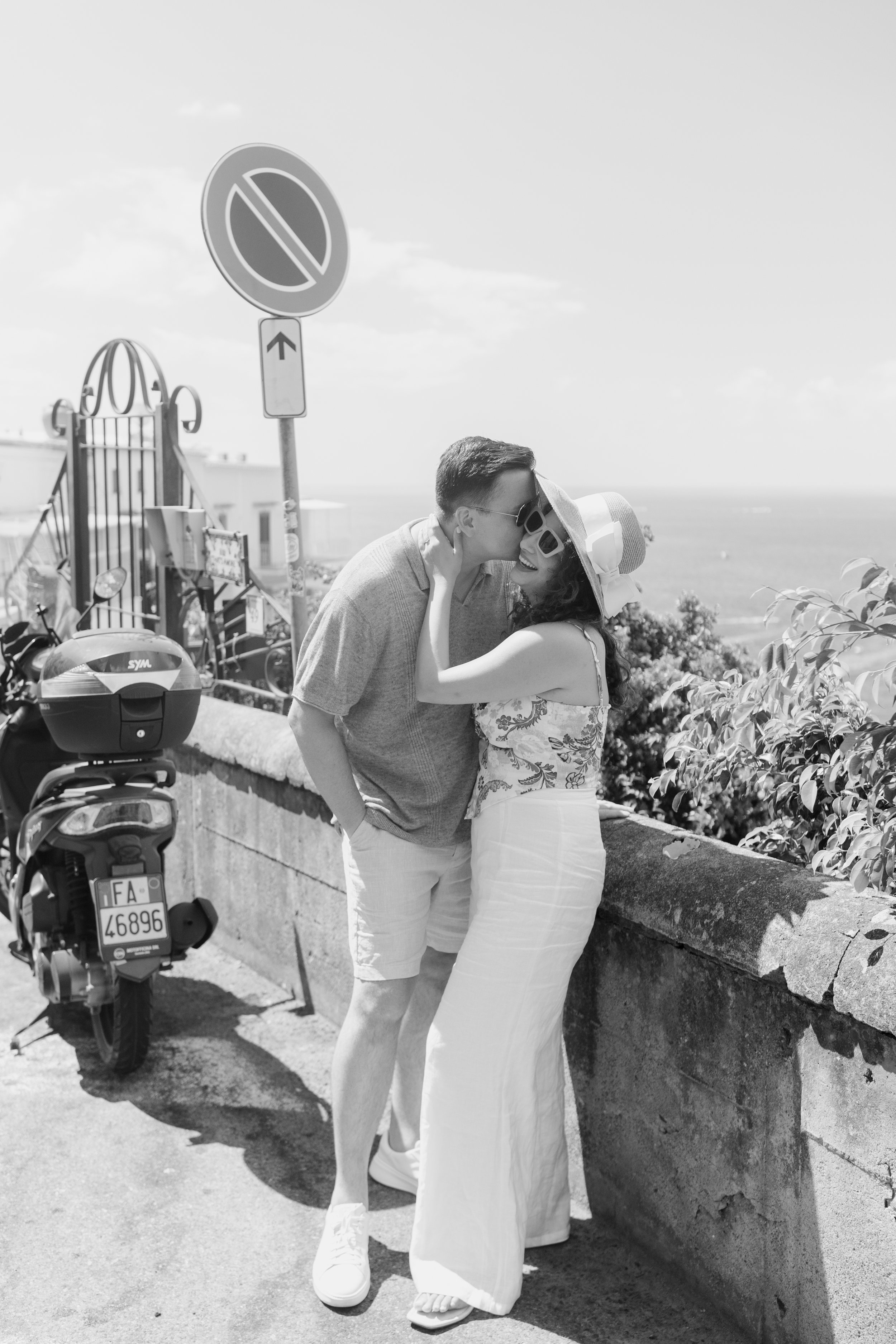 A couple sharing a kiss by a seaside outdoor area with a stone wall, street signs, and a scooter parked nearby. The man is wearing sunglasses, a light blazer, and light shorts, while the woman is dressed in a wide-brimmed hat, sunglasses, and a light