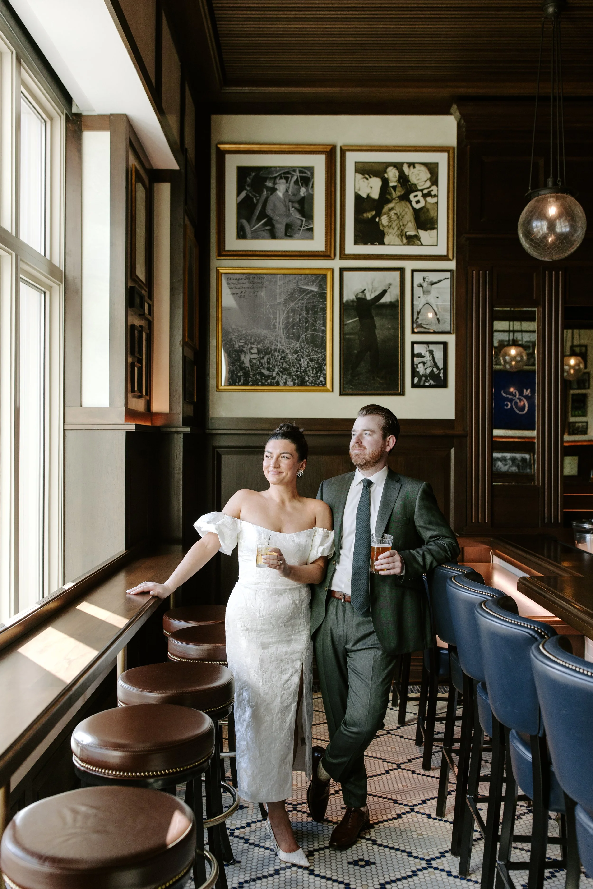 A couple dressed elegantly, the woman in a white off-shoulder dress and the man in a gray suit, standing together in a cozy, vintage-style bar with framed black-and-white photos on the wall, holding drinks, by a window with natural light, with bar st