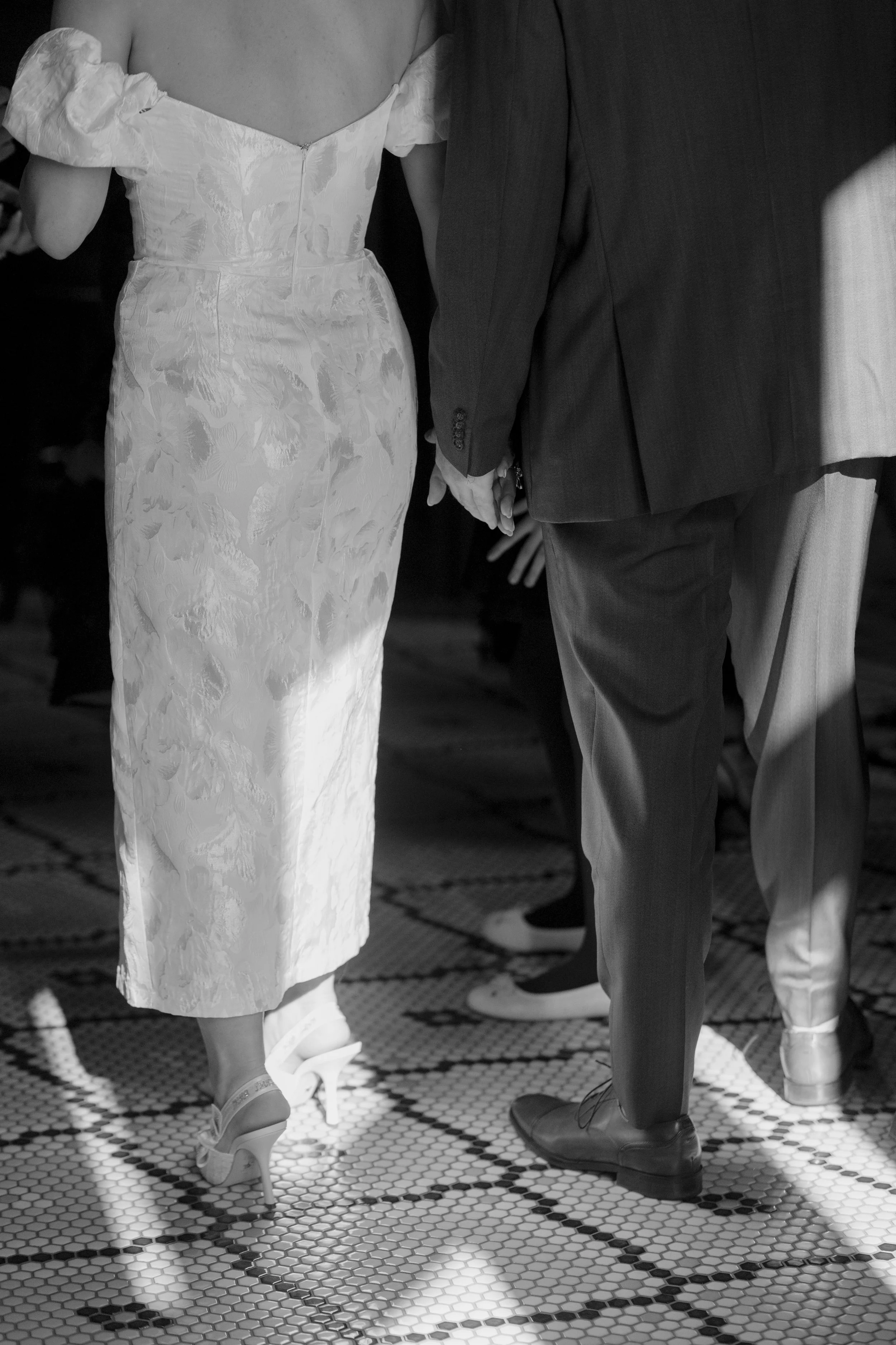 People holding hands, dressed in formal attire, standing on a tiled floor.