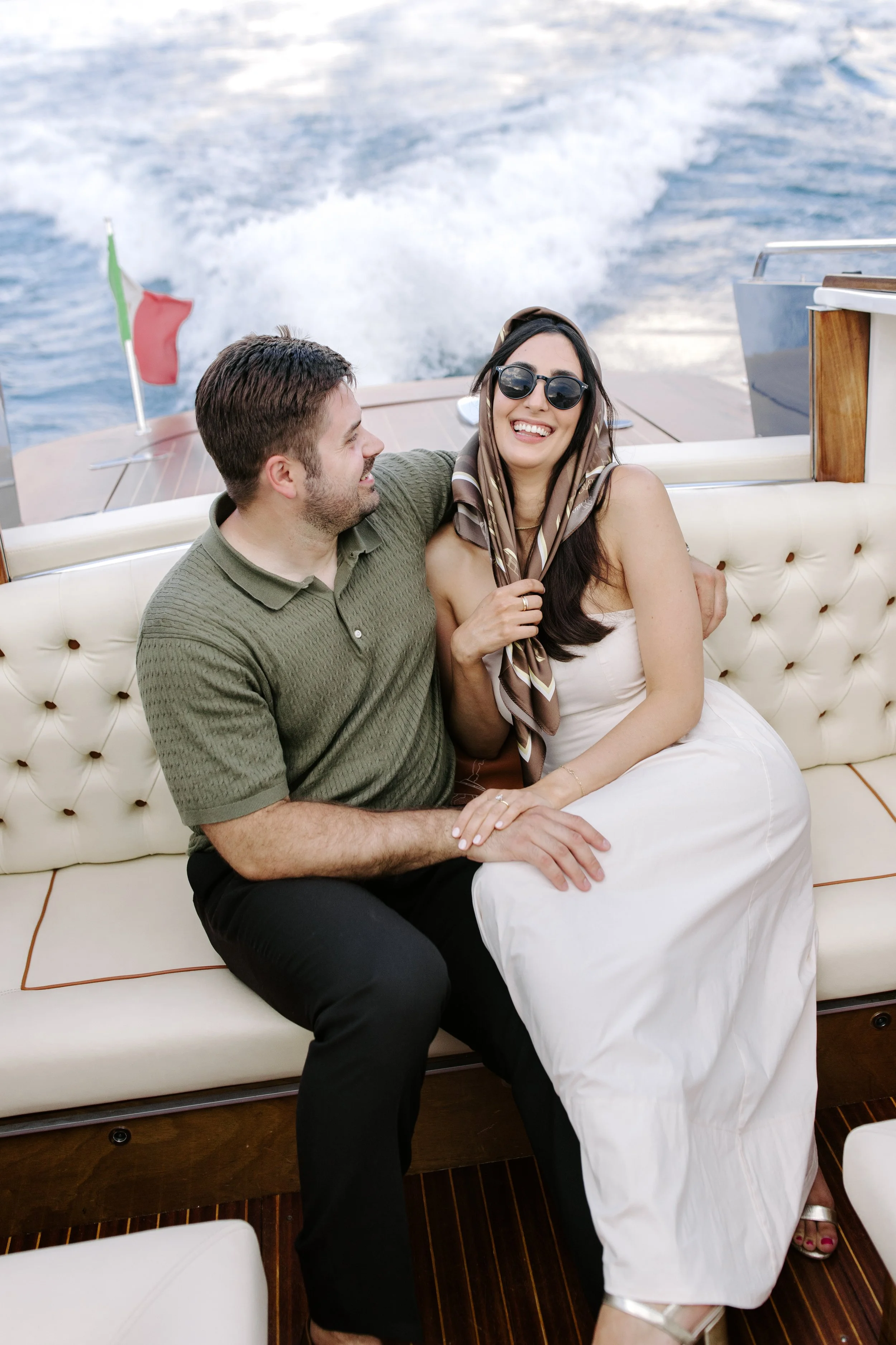 A man and woman sitting together on a boat, smiling and enjoying each other's company. The woman is wearing dark sunglasses, a headscarf, and a white dress, while the man is dressed in a green polo shirt and black pants. The background shows the ocea