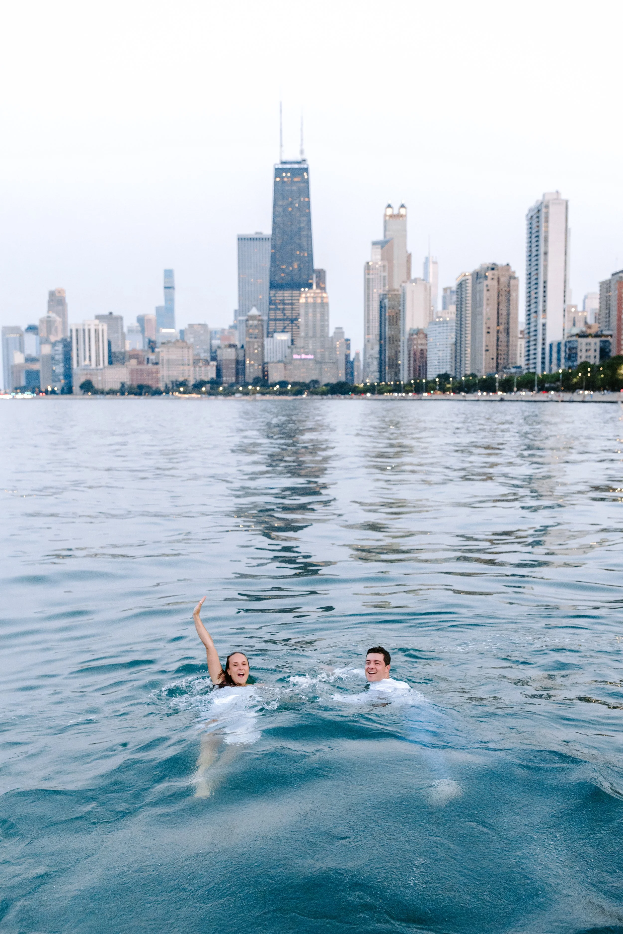 A woman and a man swimming in a body of water with a city skyline in the background.