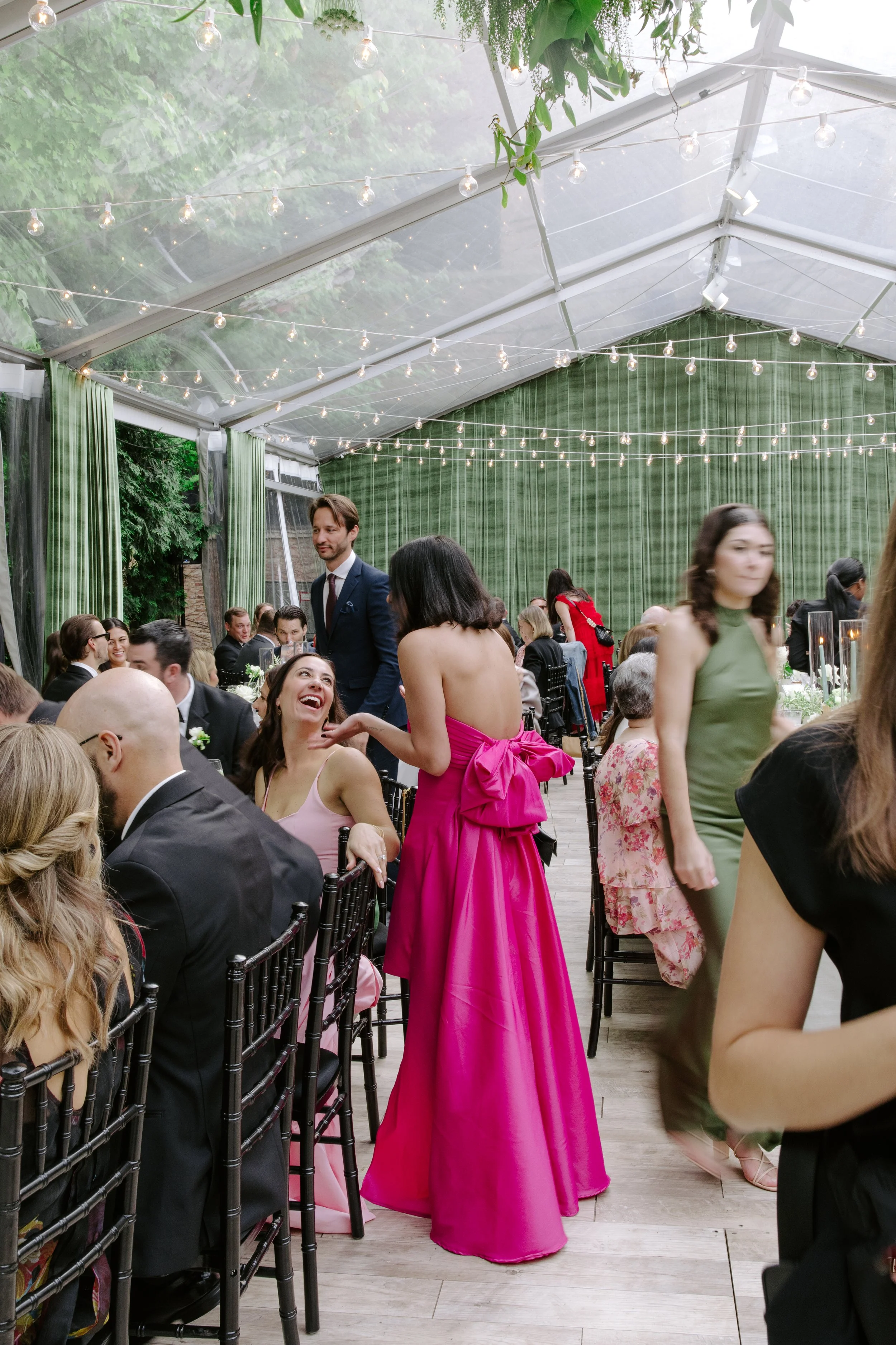 People at a formal gathering or wedding reception inside a large tent with string lights, green curtains, and a wooden floor. Some individuals are seated at tables, engaged in conversation, while others are standing or walking.
