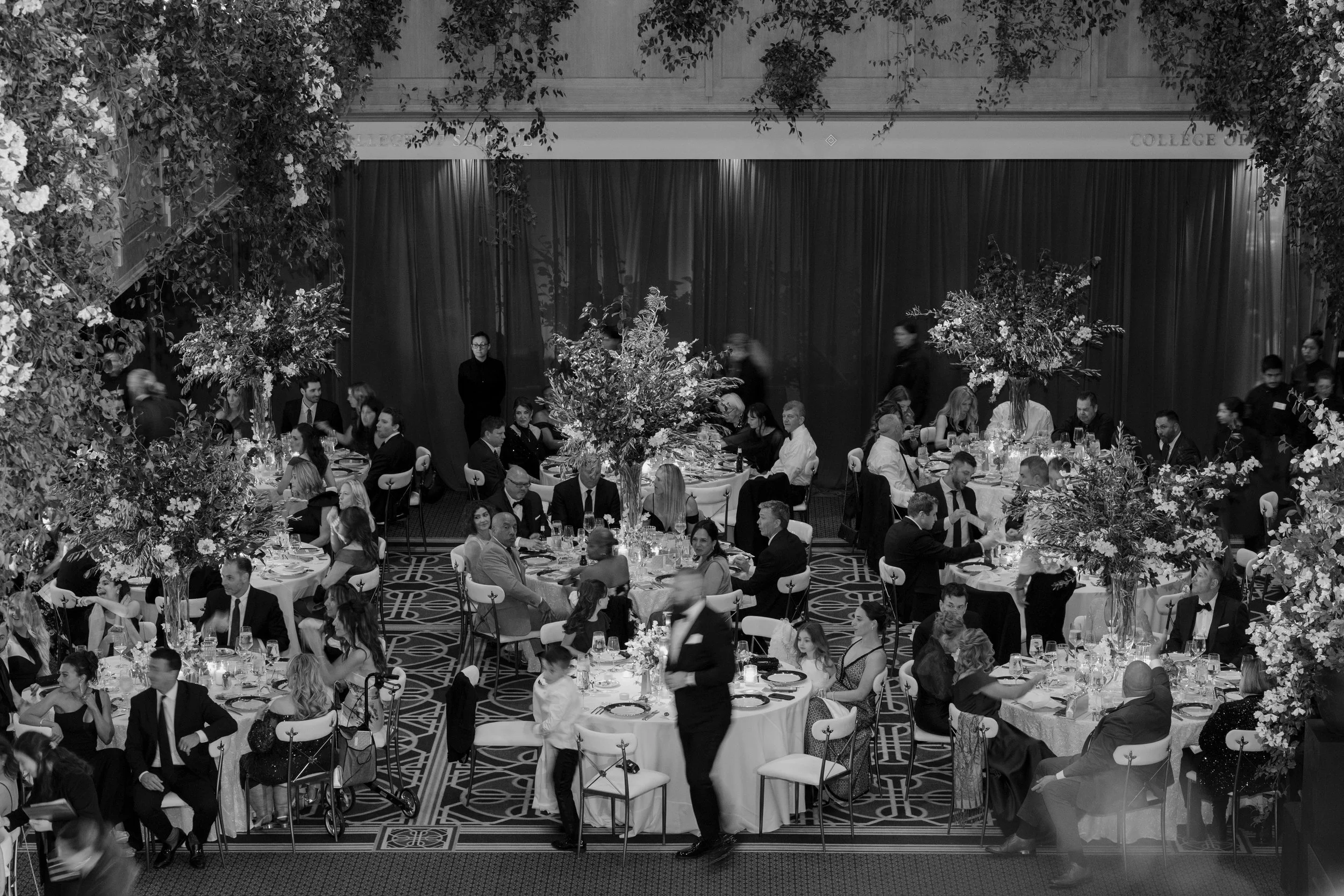 A large banquet hall with many round tables set for dining, filled with formally dressed guests. The room is decorated with tall floral centerpieces and surrounded by dense foliage and flowers. People are sitting, eating, and talking, with some waits