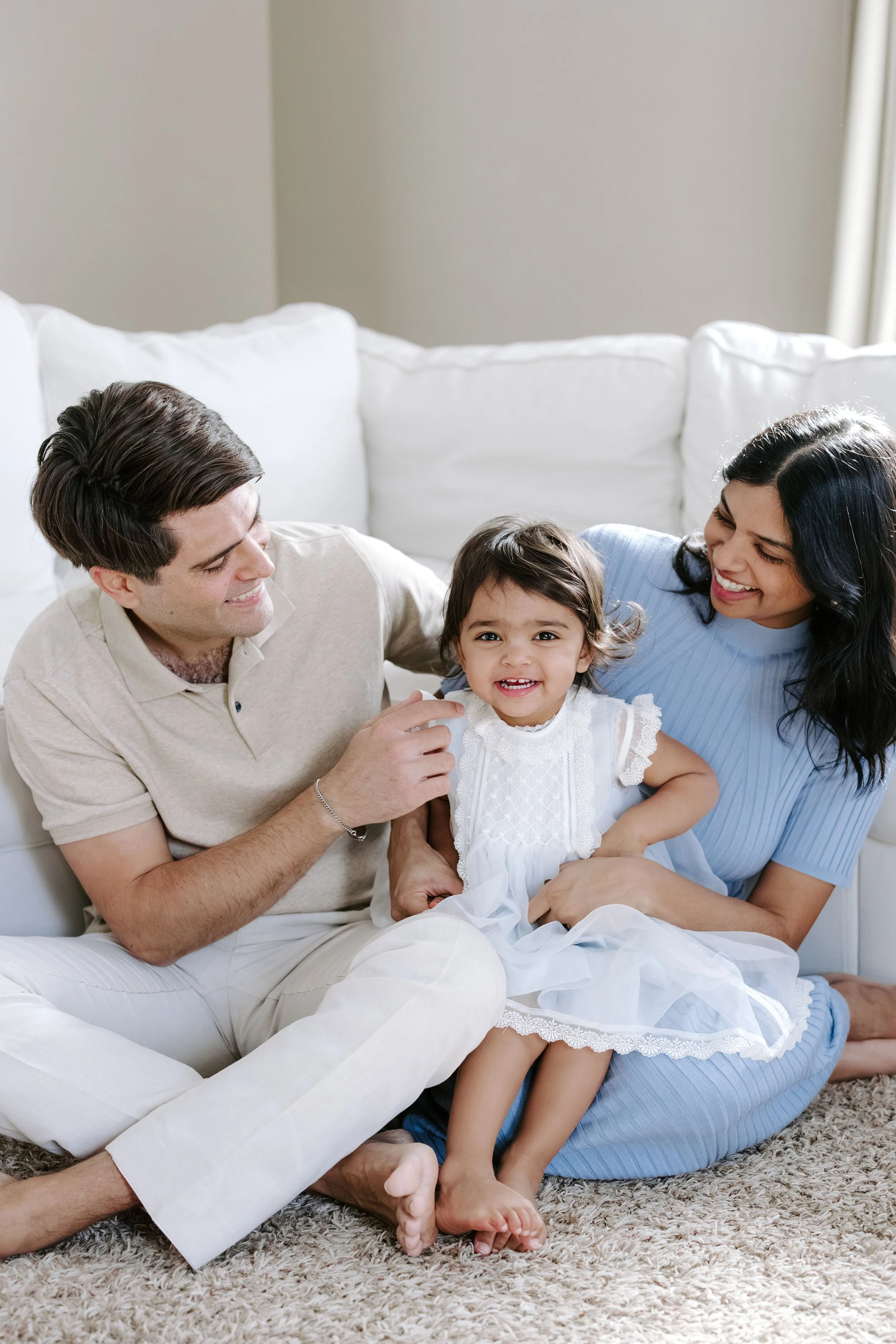 A family of three, including a man, a woman, and a young girl, sitting on a carpeted floor in a living room, smiling and enjoying each other's company.