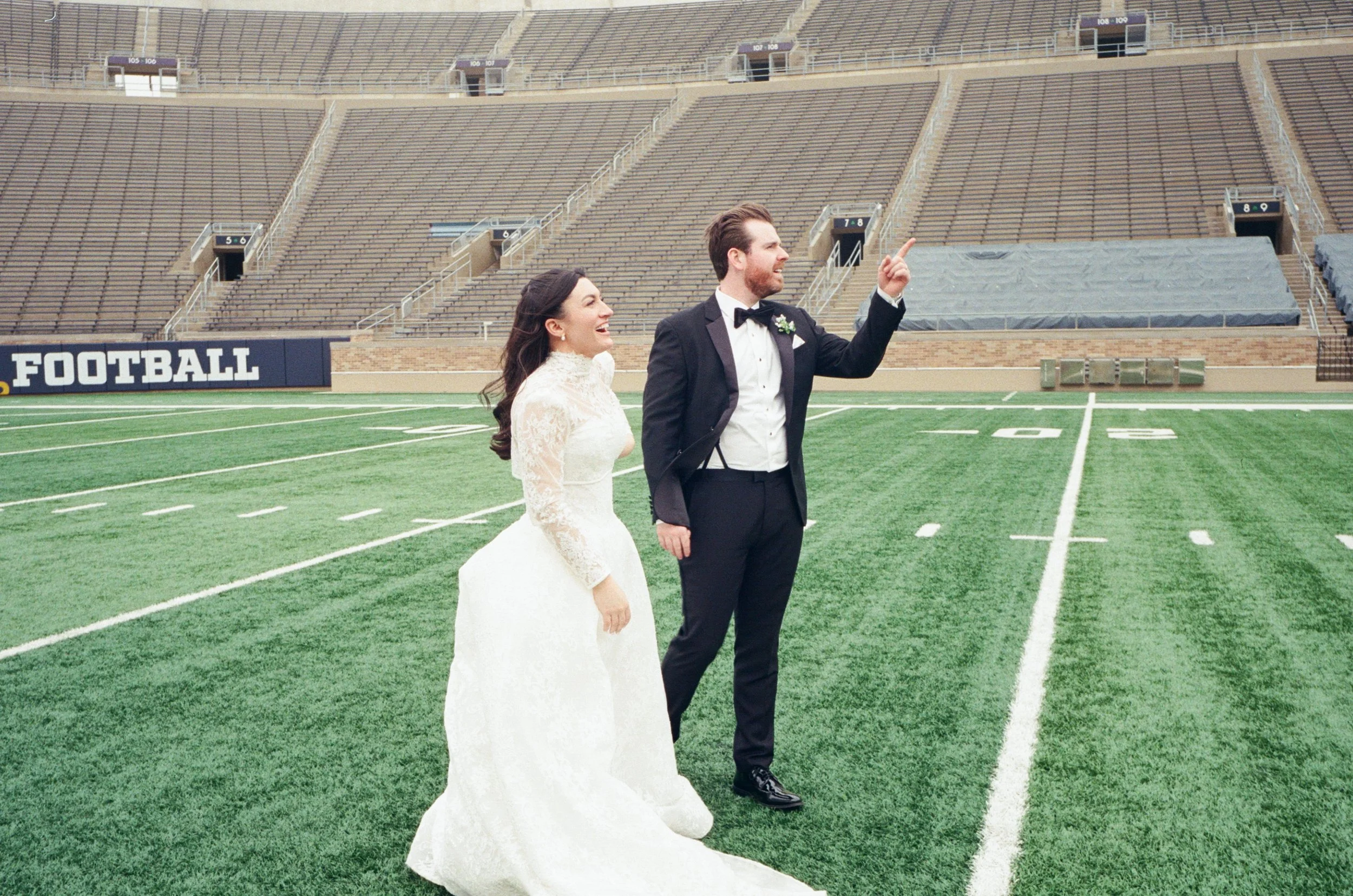 A bride and groom standing on a football field in a stadium, smiling and enjoying their wedding day.