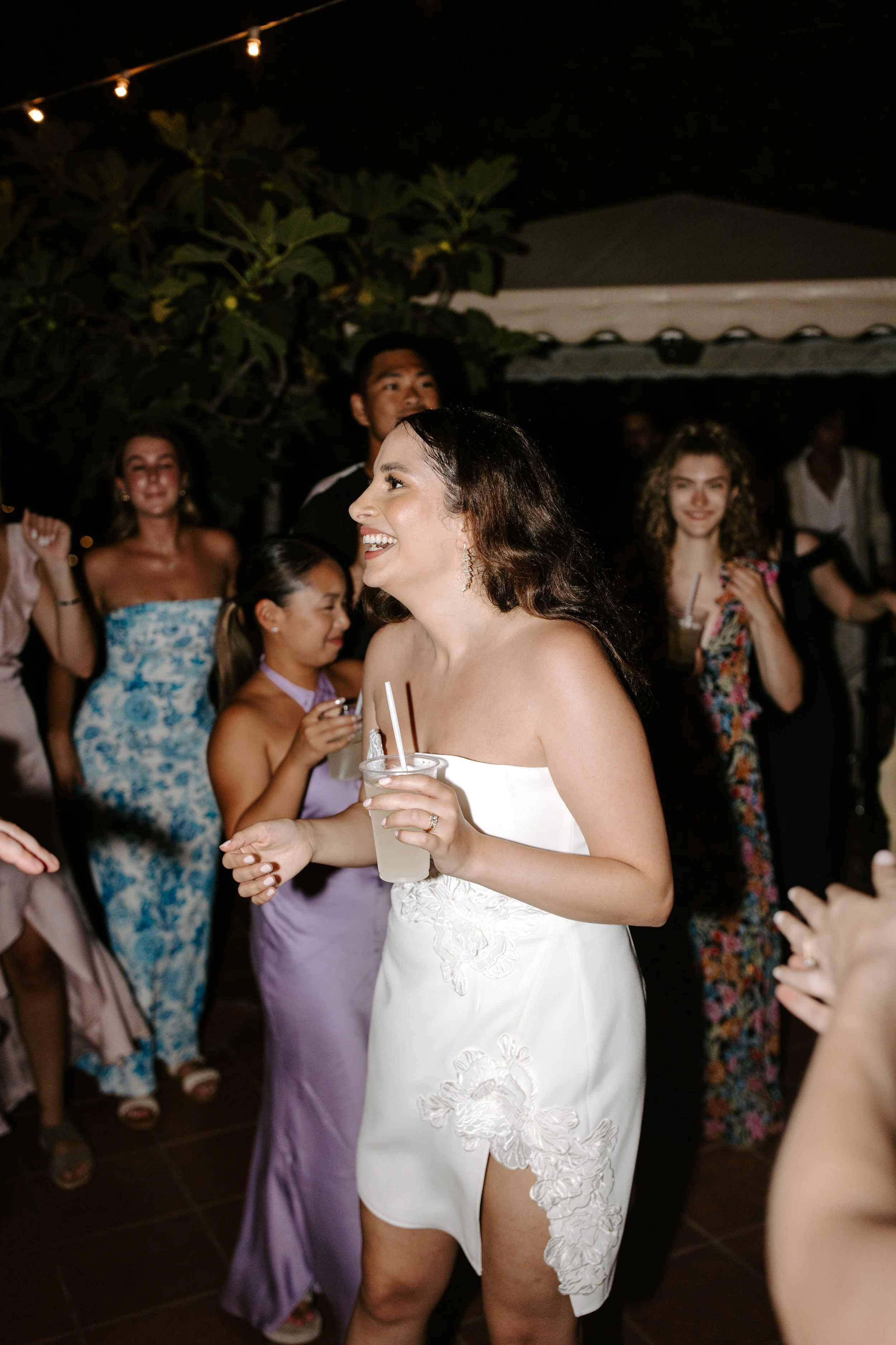 A woman in a white dress holding a drink, smiling, at a nighttime celebration with other women in colorful and floral dresses, outdoor setting with string lights overhead.