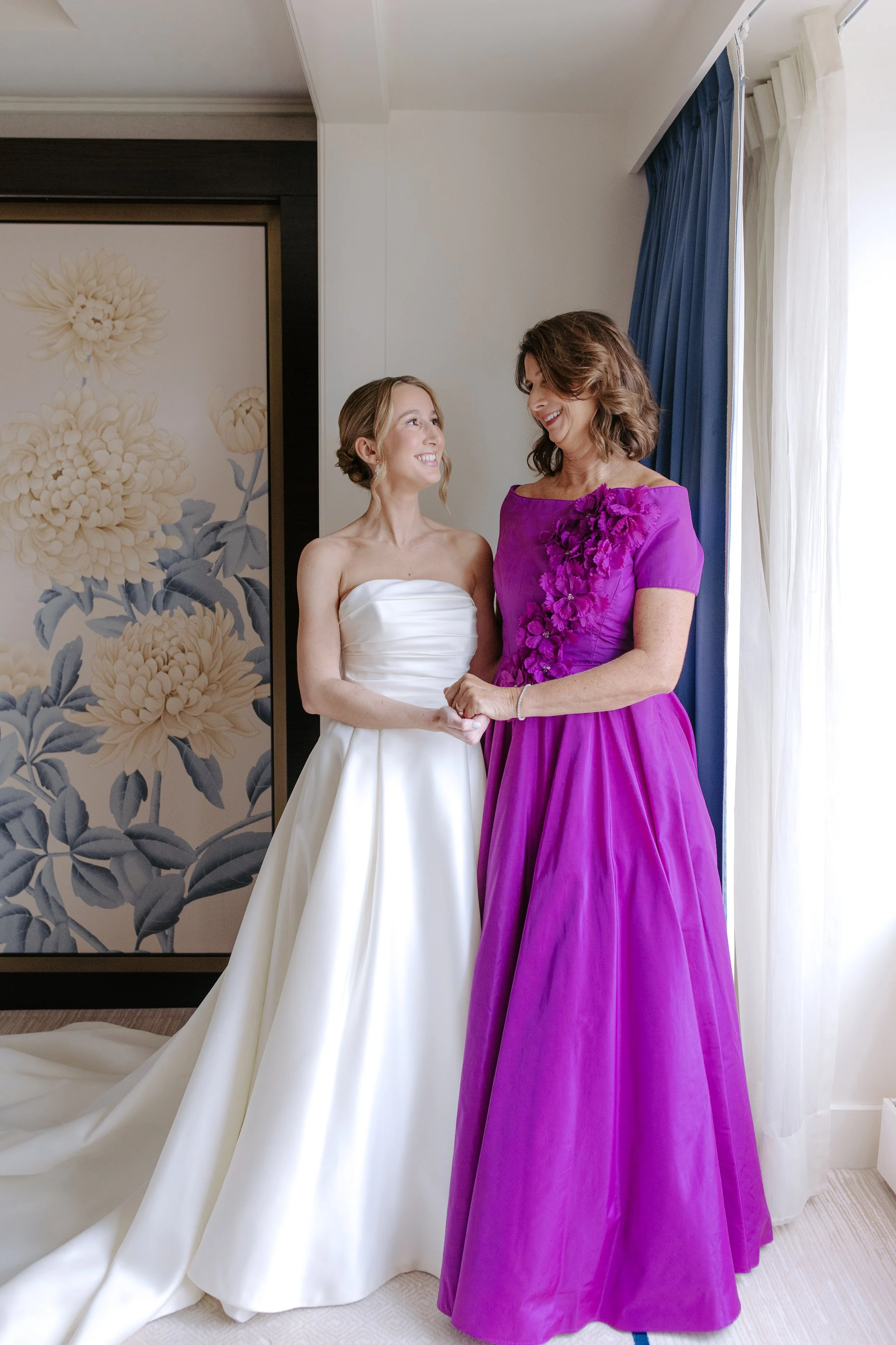 A bride in a white strapless wedding gown holding hands with a woman in a purple gown with floral embellishments, smiling and looking at each other, indoors near a window with blue curtains and beige drapes, with a floral patterned wall in the backgr
