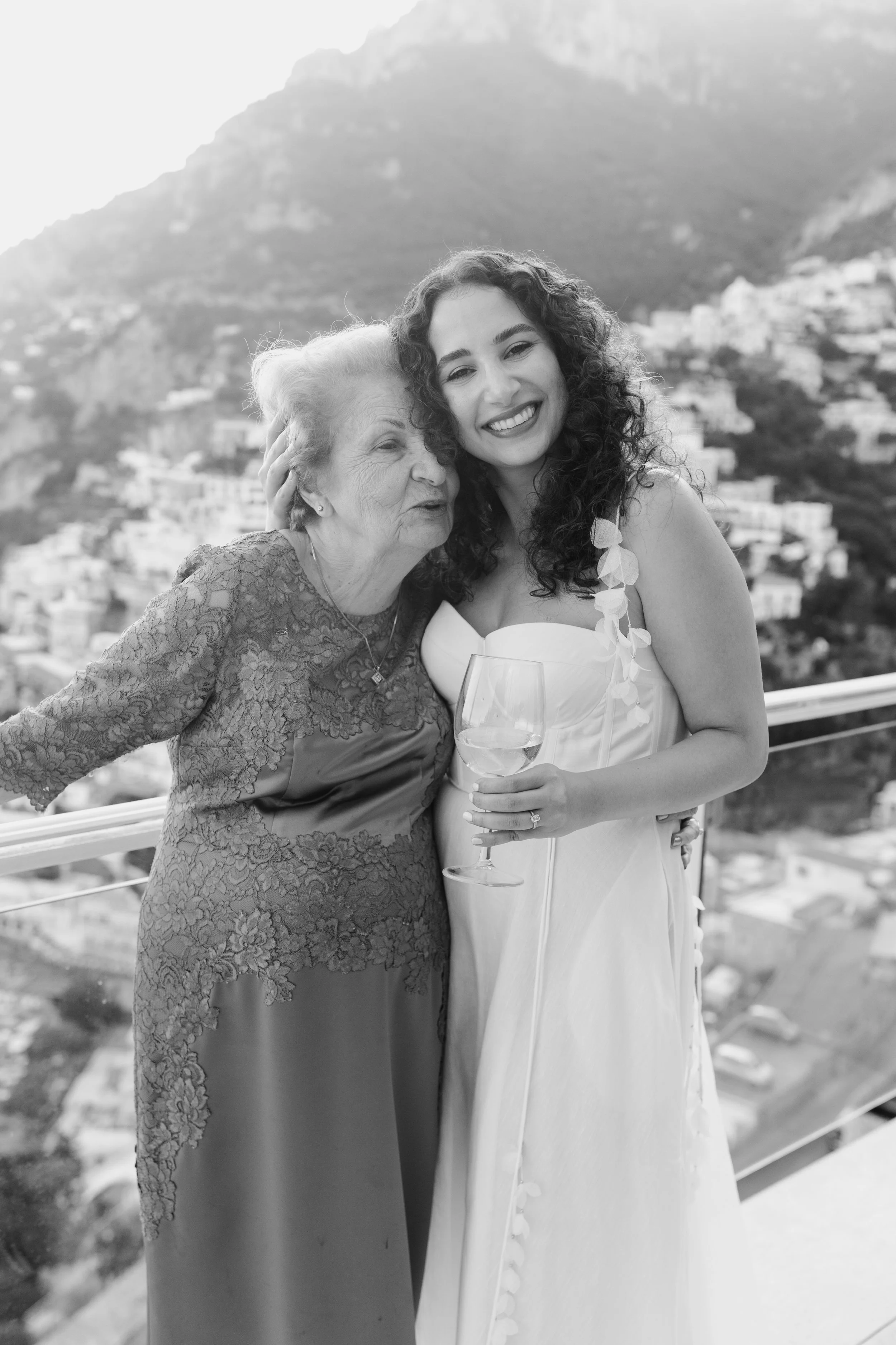 A young woman in a wedding dress and an older woman in a lace dress smiling and hugging each other outdoors with a mountainous town in the background.