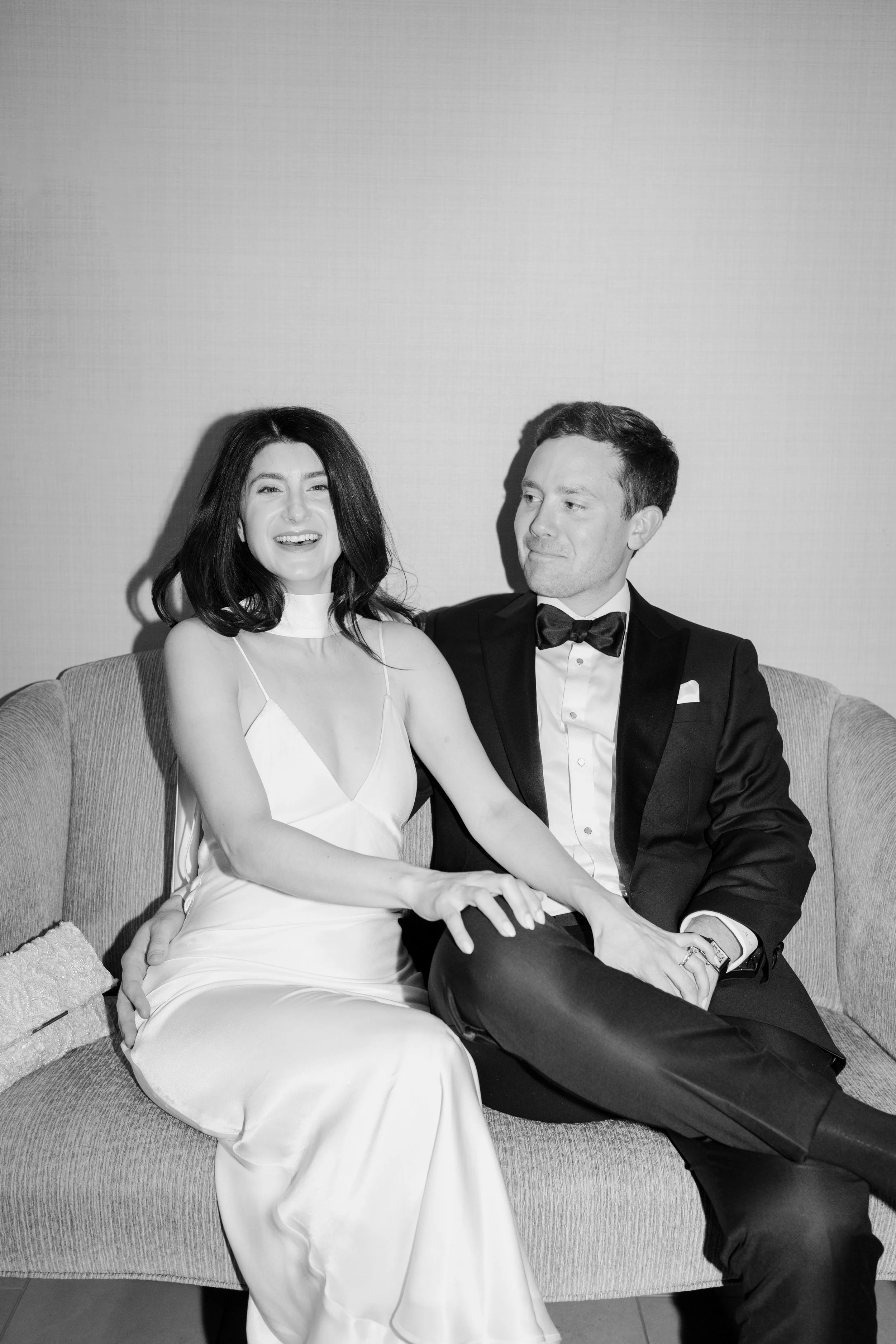 A black and white photo of a smiling woman in a white dress and a man in a tuxedo sitting on a sofa, holding hands and looking at each other.