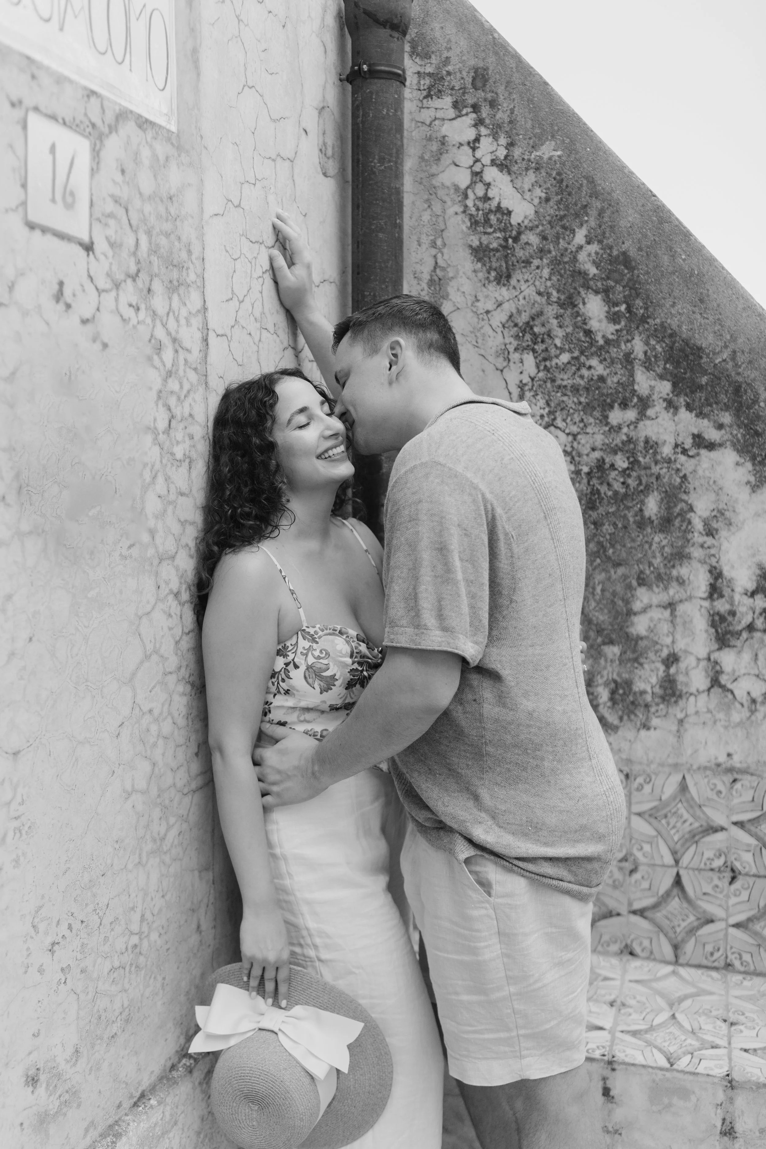 A couple shares an intimate moment in a black-and-white photo; they are close, smiling, and about to kiss, with the woman leaning against a textured wall and the man facing her with his hand on the wall.