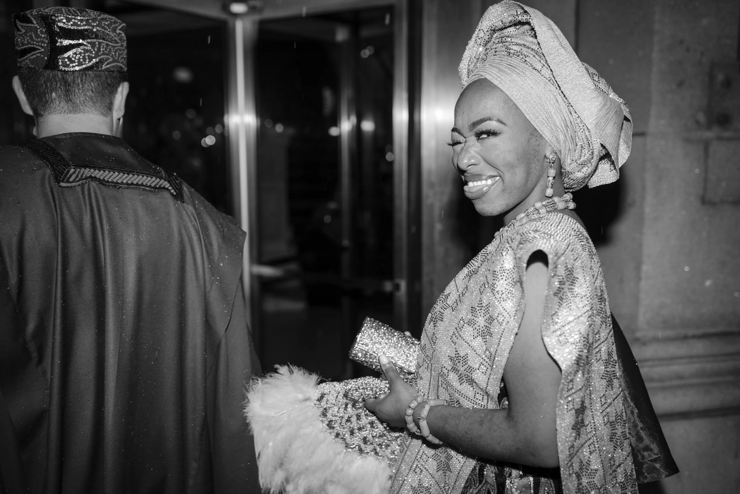 Black and white photo of a woman in traditional African attire, smiling and winking. She is wearing an ornate head wrap, jewelry, and a sequined dress, holding a clutch and wearing a bracelet, with her hand adorned with fur details.
