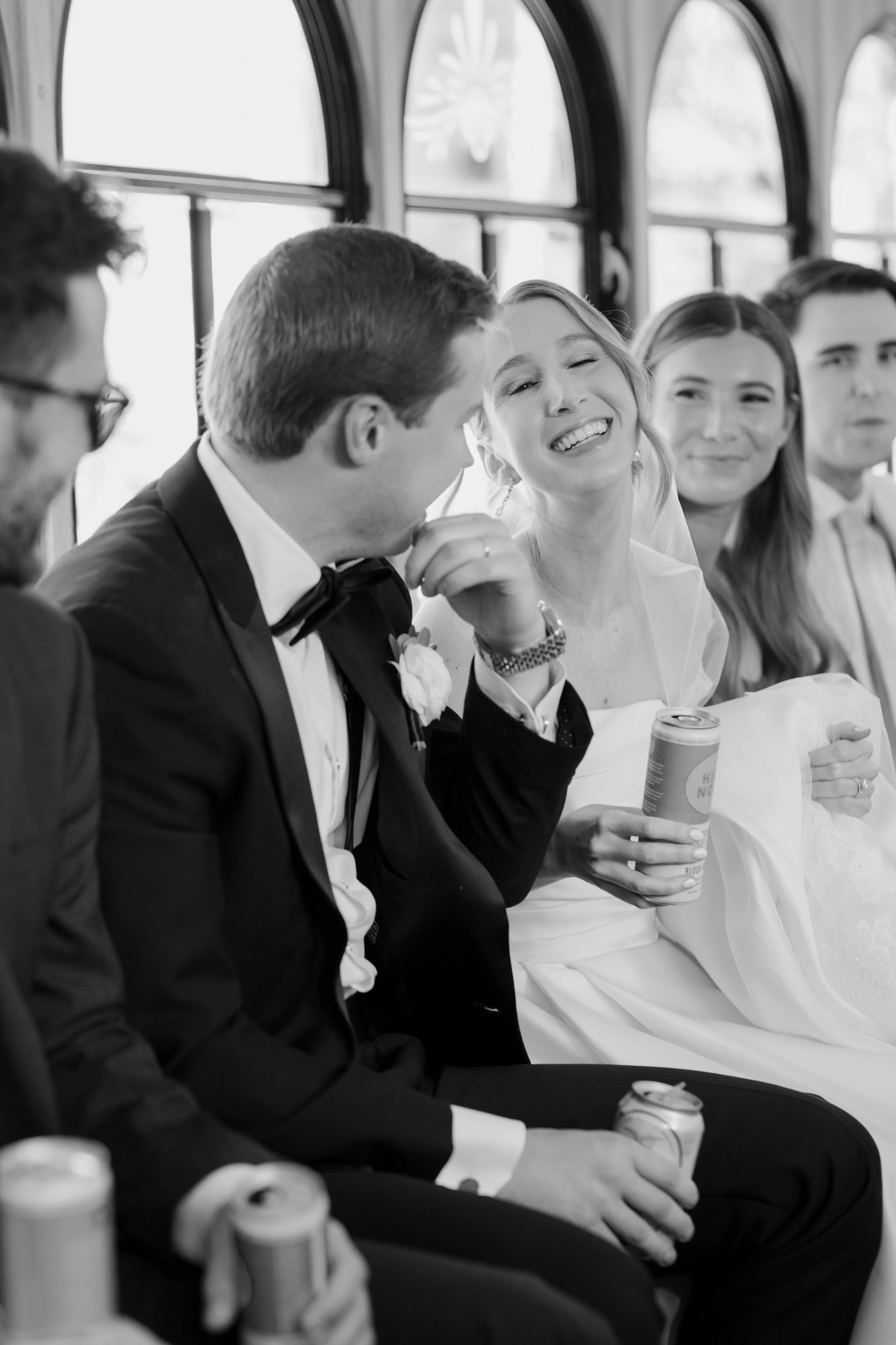 Black and white photo of people in formal attire sitting in a row, smiling, with drinks in their hands, inside a bus or train with arched windows and decorative elements on the windows.