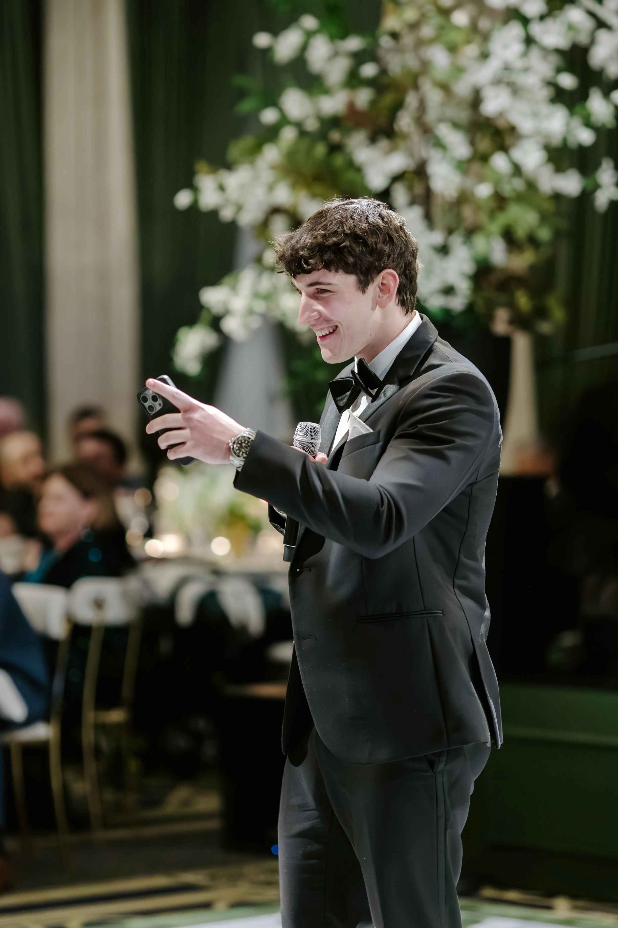 Young man dressed in a black tuxedo with a bow tie, smiling, holding a microphone and a smartphone, at a formal event with floral decorations and seated guests in the background.