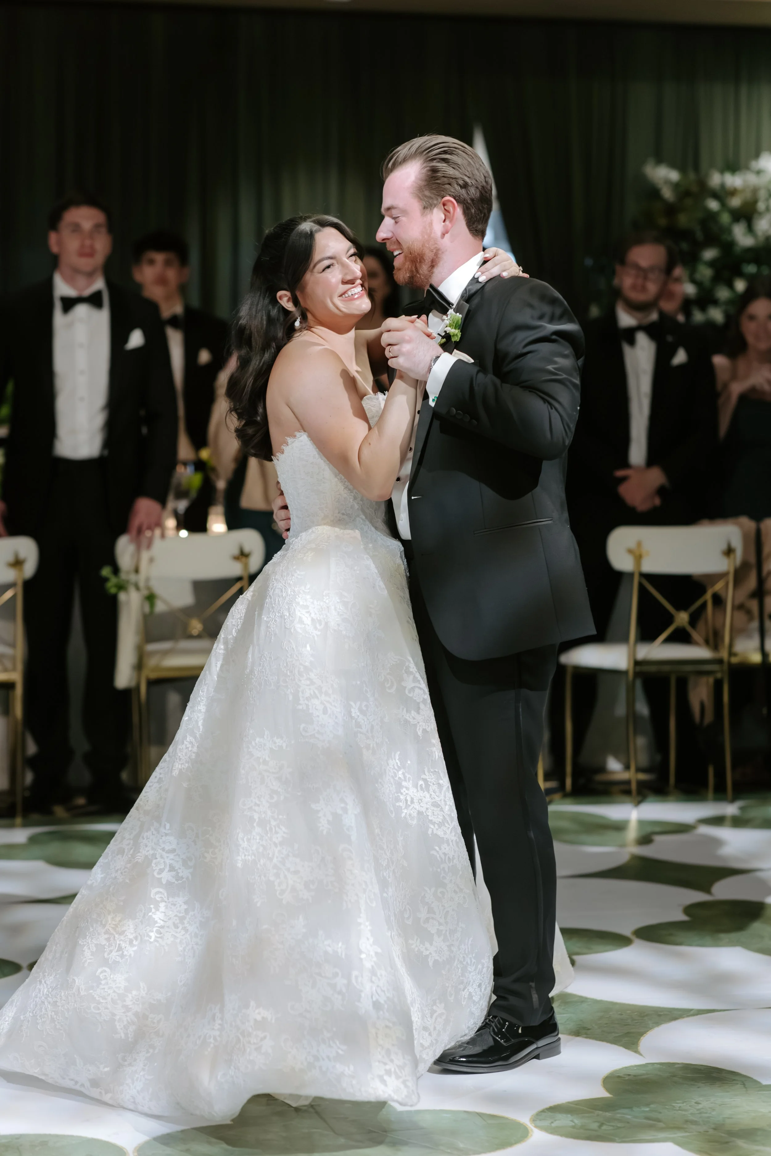 A bride and groom are dancing during their wedding reception, smiling and looking at each other, with wedding guests in formal attire in the background.
