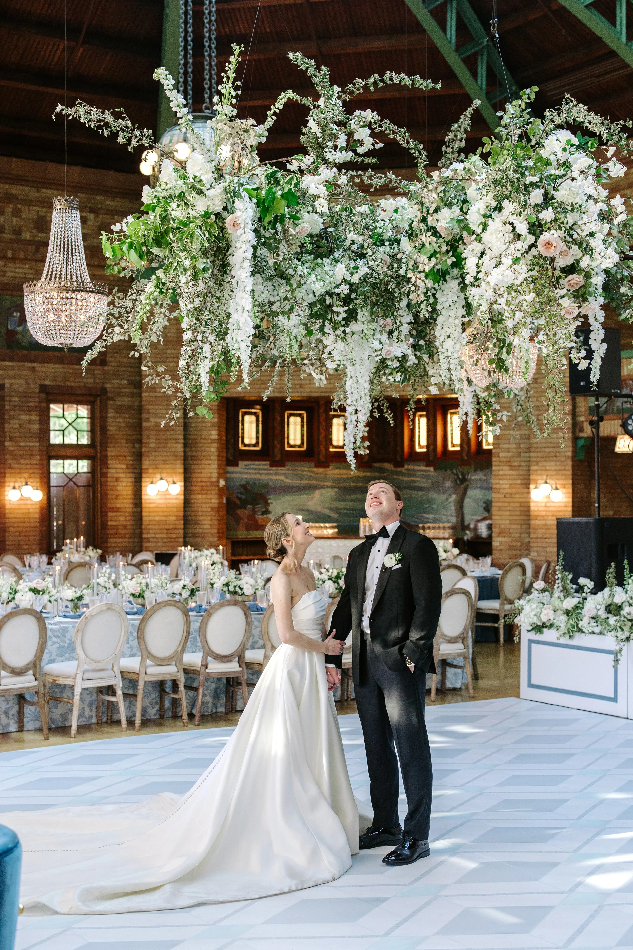 A bride and groom holding hands and smiling at each other in a decorated wedding venue with a large floral arrangement hanging from the ceiling.