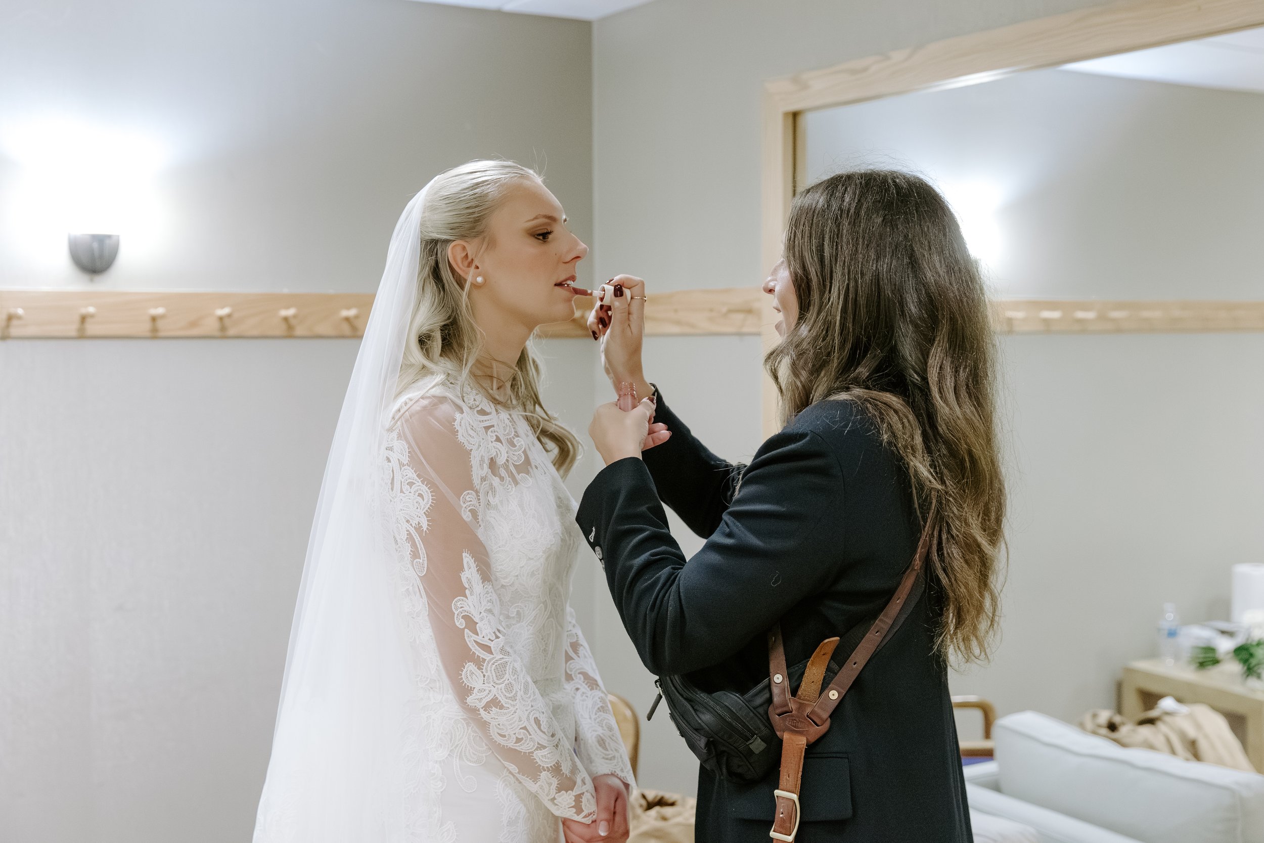 A bride with blonde hair in a lace wedding dress is getting ready, assisted by a woman with dark hair in a black jacket, in a room with light-colored walls and a mirror.