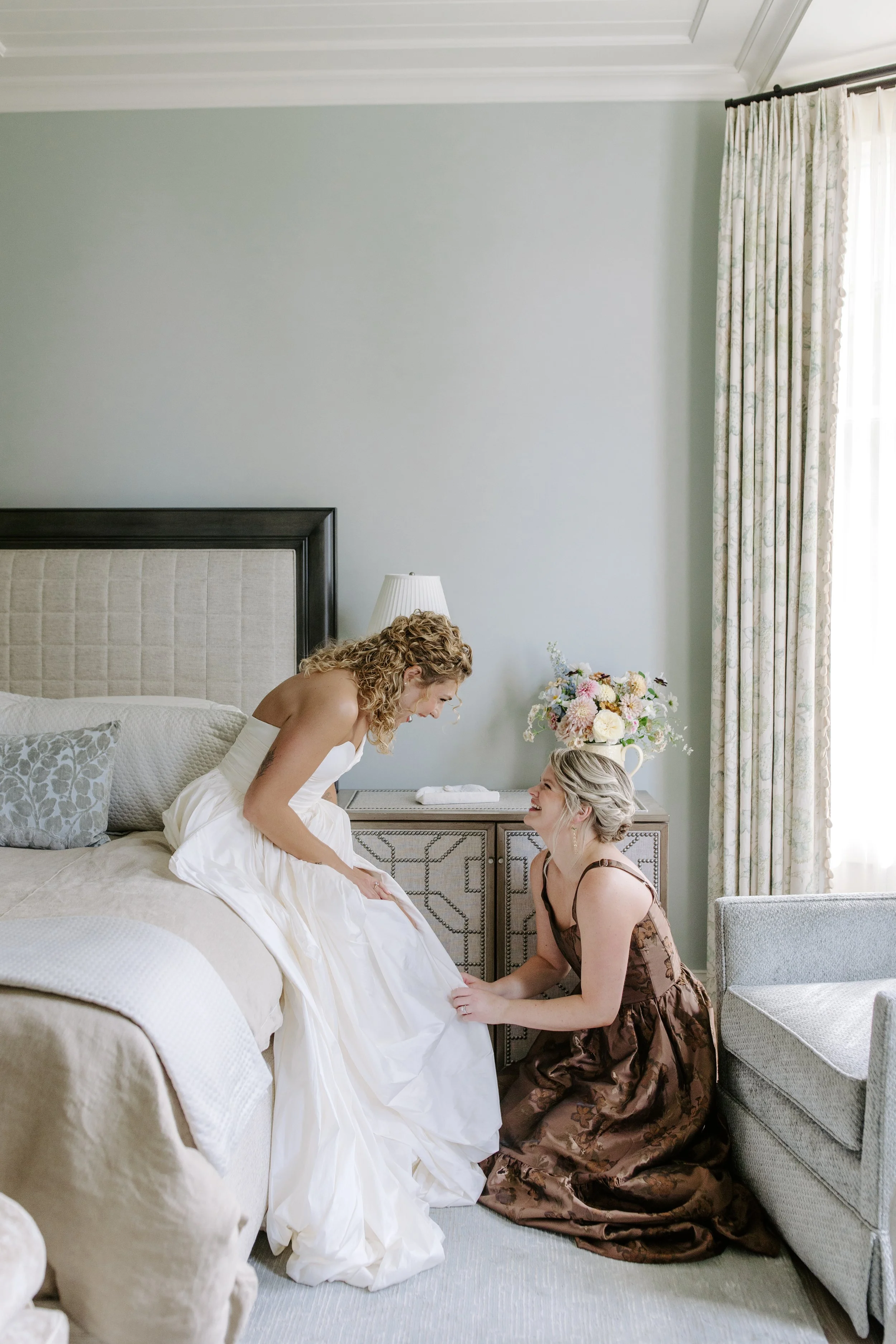 Two women are in a bedroom. One is sitting on a bed, wearing a white dress, and the other is kneeling in front of her, wearing a brown dress. They are smiling and looking at each other. There is a nightstand with a vase of flowers, a lamp, and some t