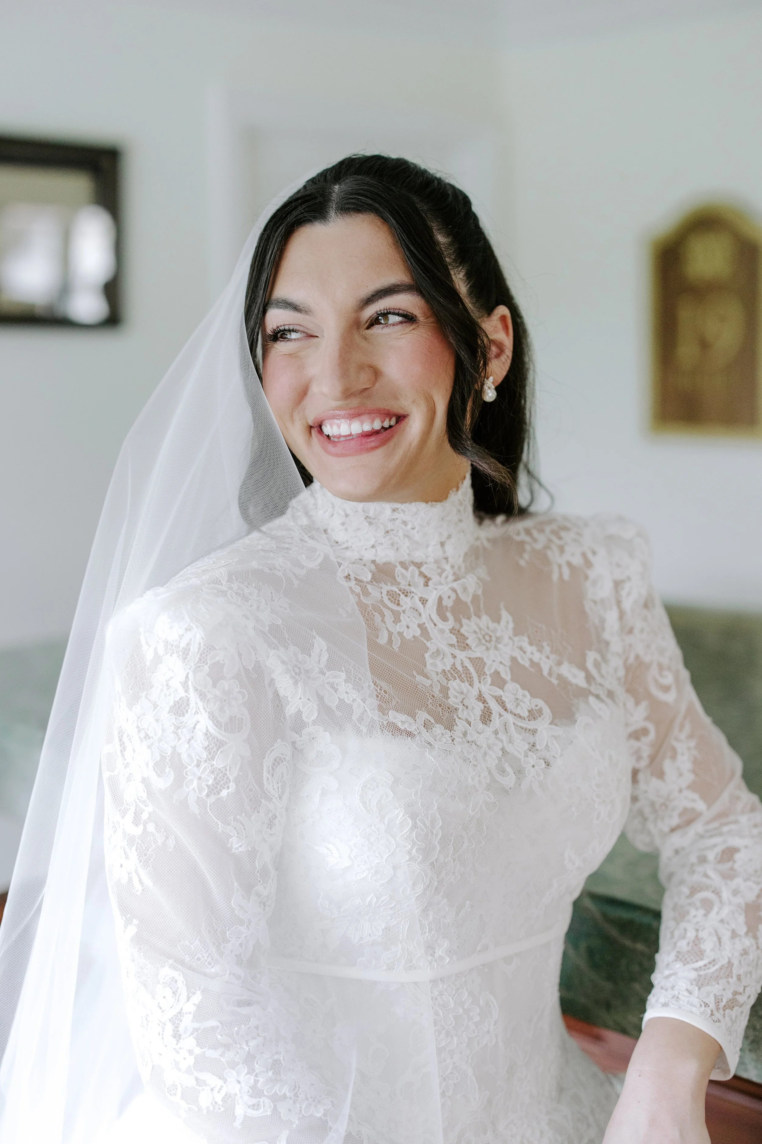 A smiling bride wearing a white lace wedding dress with a high neck, sheer floral lace overlay, and long sheer sleeves. She has dark hair styled in loose waves and is wearing pearl earrings, with a white veil draped over her head.