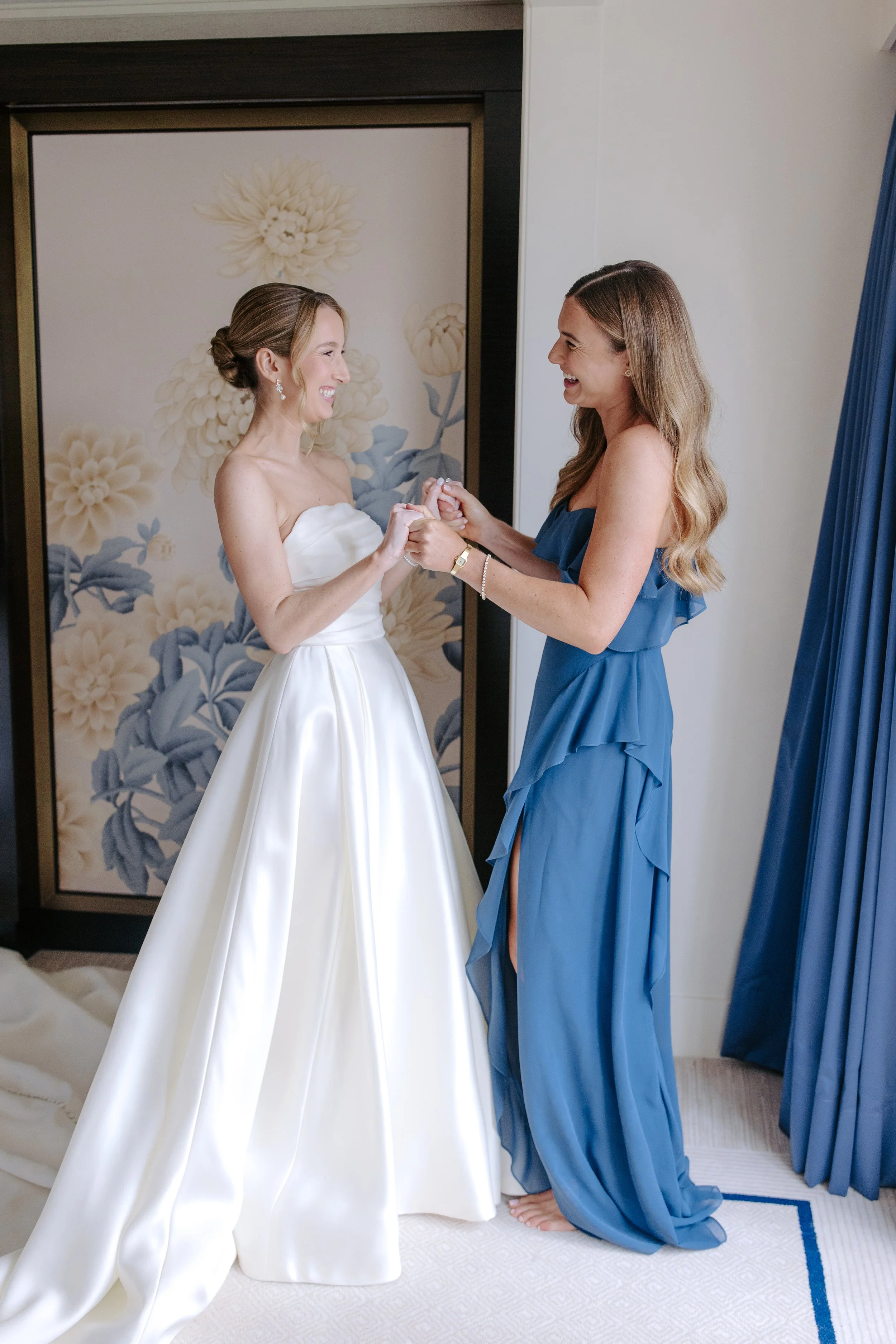 A bride in a white strapless wedding gown holding hands with a woman in a blue dress, both smiling at each other inside a room with floral decor and blue curtains.