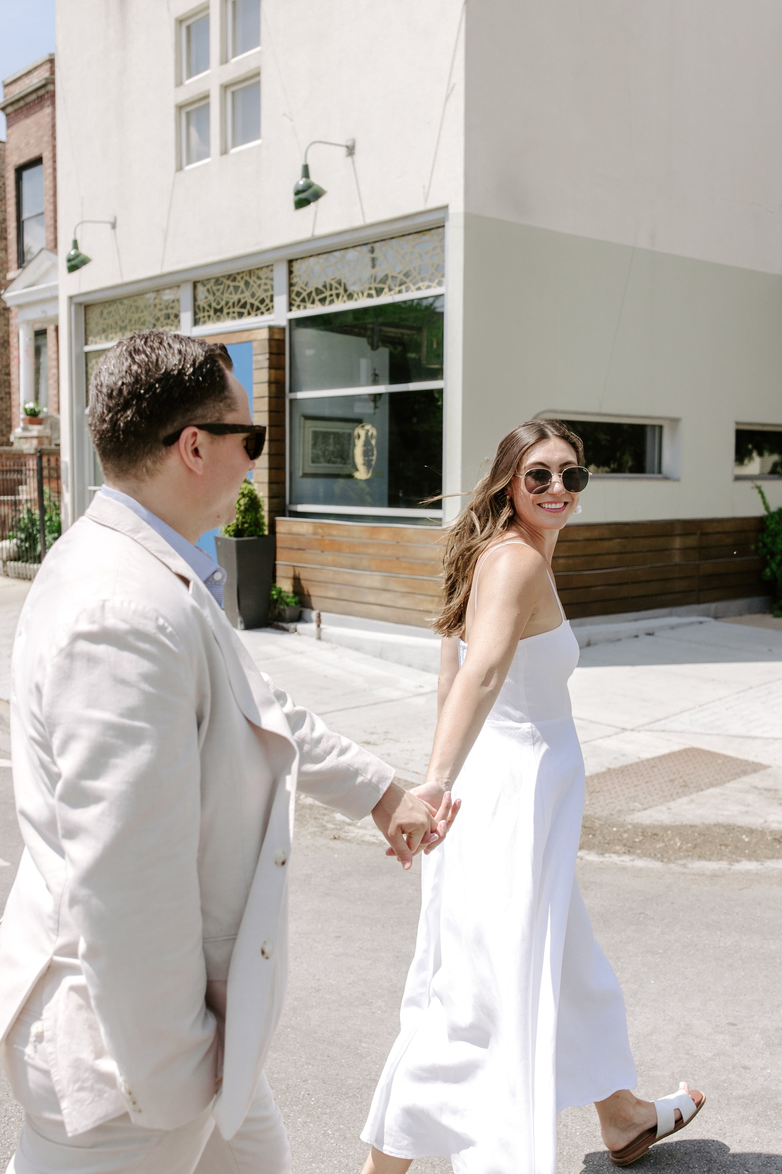 A man and woman holding hands and walking on the street in bright sunlight. The woman is smiling and wearing sunglasses and a white dress, the man is wearing a white suit and sunglasses.