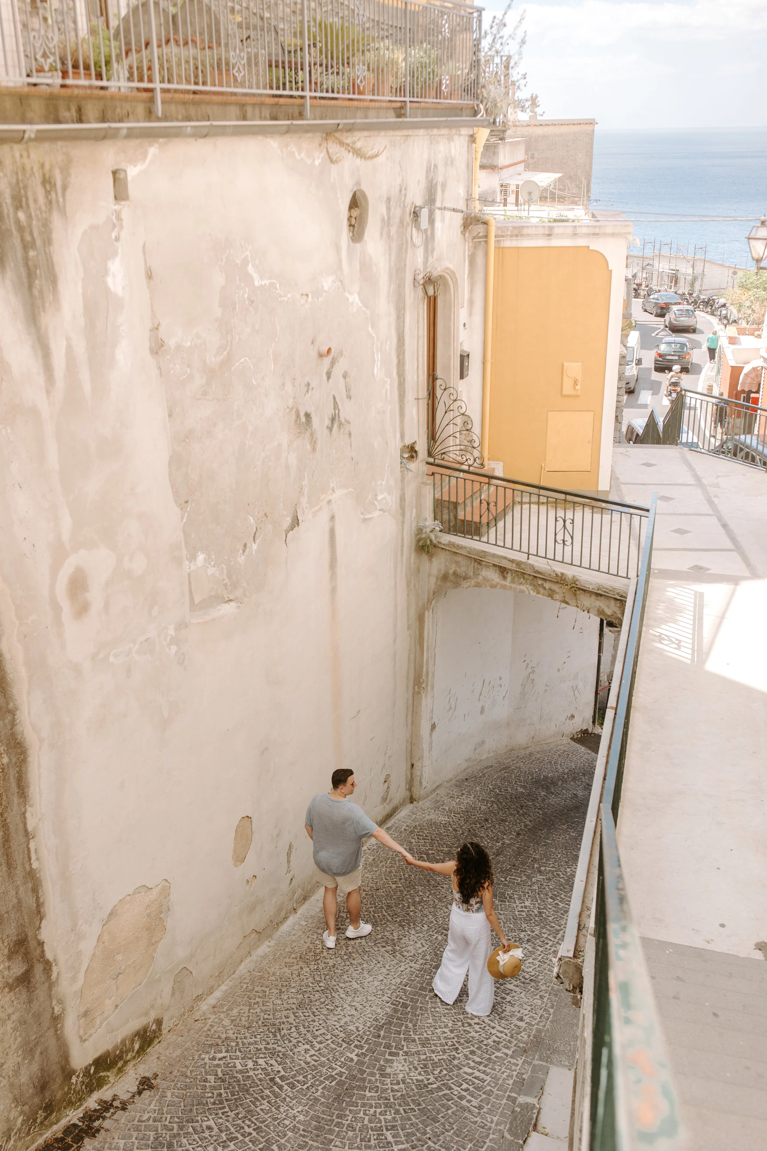 A man and a woman holding hands walking down a curved cobblestone street in a European seaside town, with a yellow building and a view of the ocean in the background.