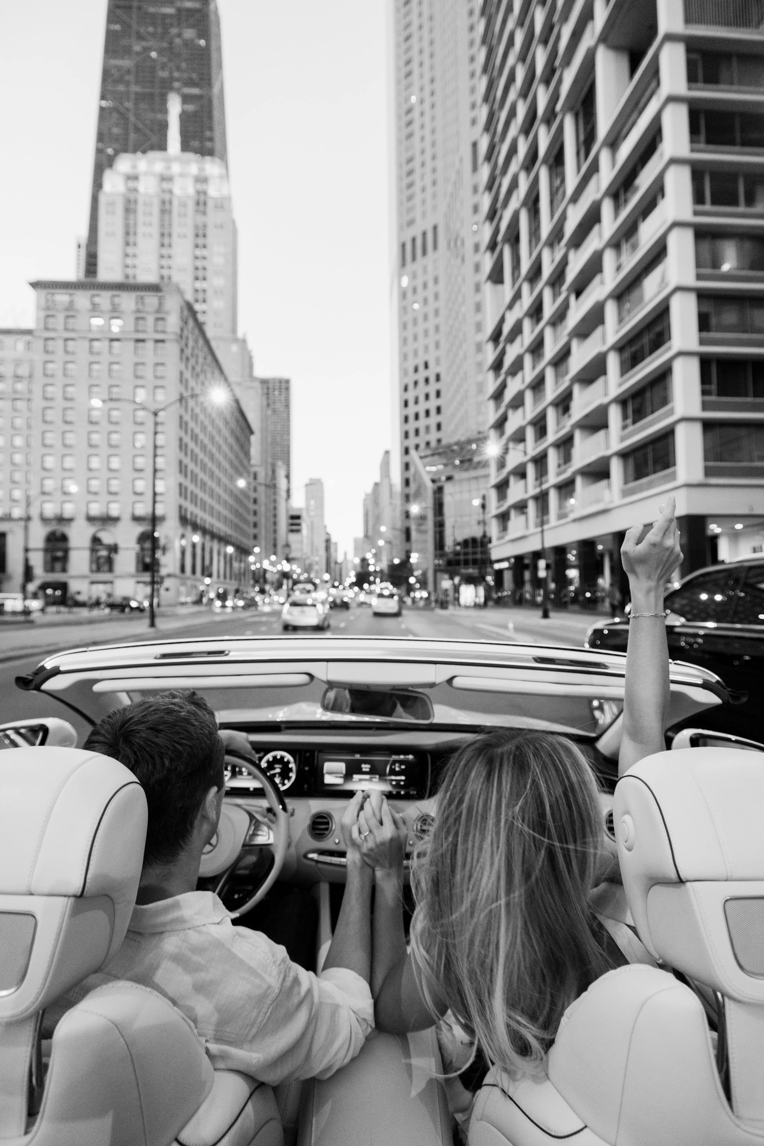 View from inside a convertible car with three passengers, showing a city street with tall buildings and streetlights, and one person raising their arm.