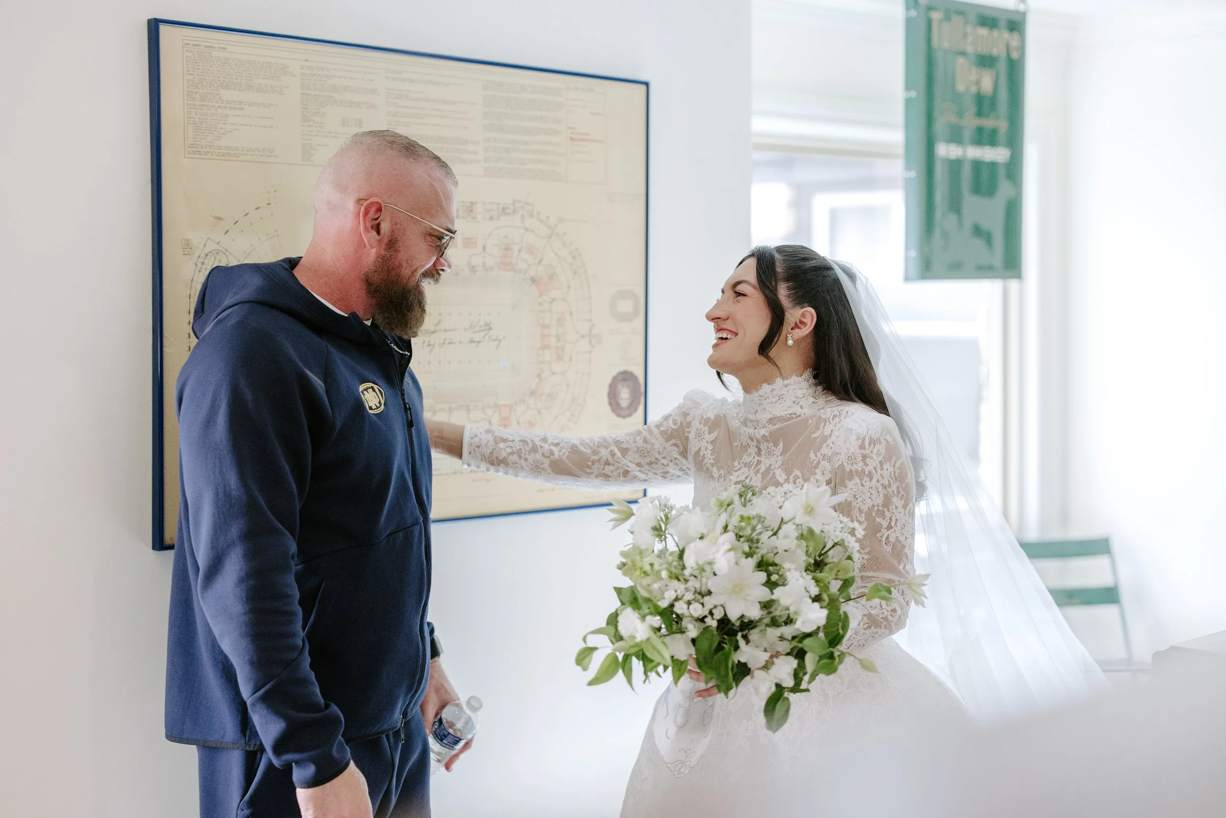 A bride in a lace wedding dress holding a bouquet of white flowers and smiling at a man in a navy jacket with a badge, standing against a wall with a large map or diagram, during a wedding ceremony.