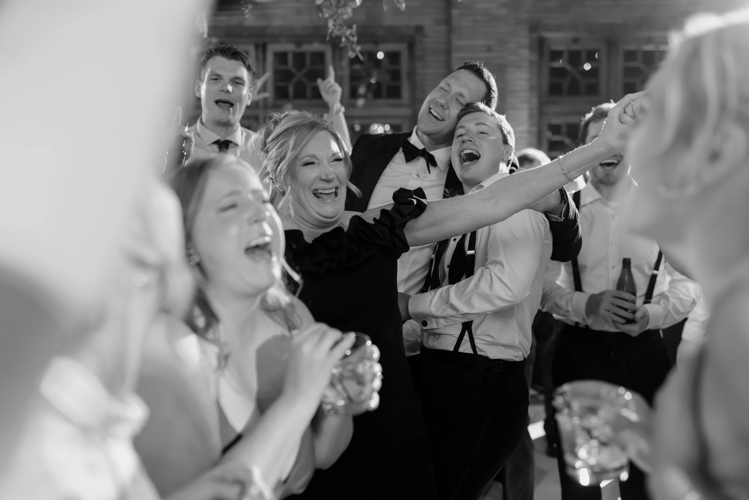 People dressed in formal attire at a social gathering, laughing and enjoying themselves, with some holding drinks, in a rustic wooden venue.