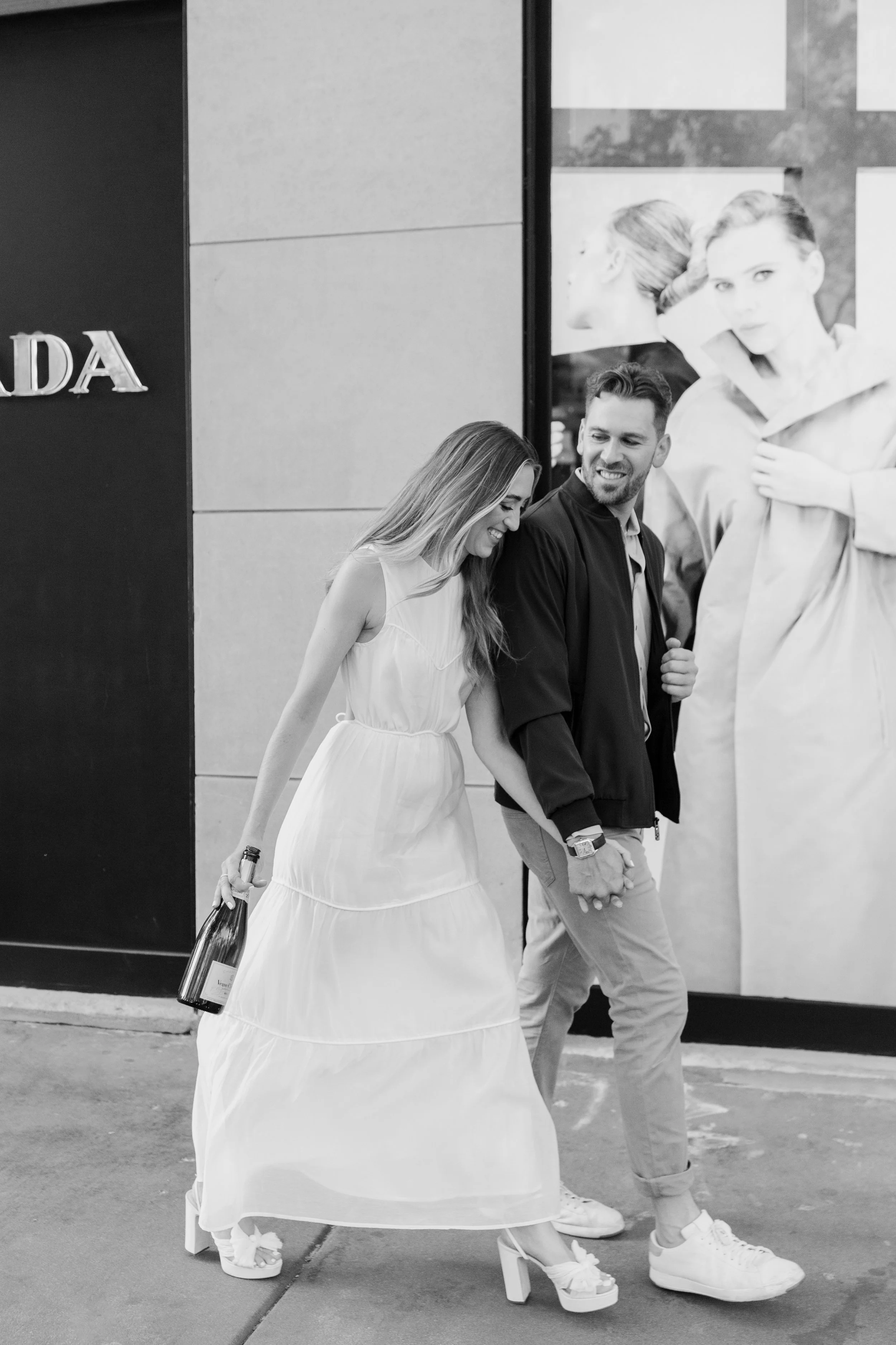 A black-and-white photo of a man and woman walking hand-in-hand on the street. The woman is wearing a long white dress and high heels, holding a bottle of wine, and smiling. The man is dressed casually in a jacket, pants, and sneakers, and is smiling