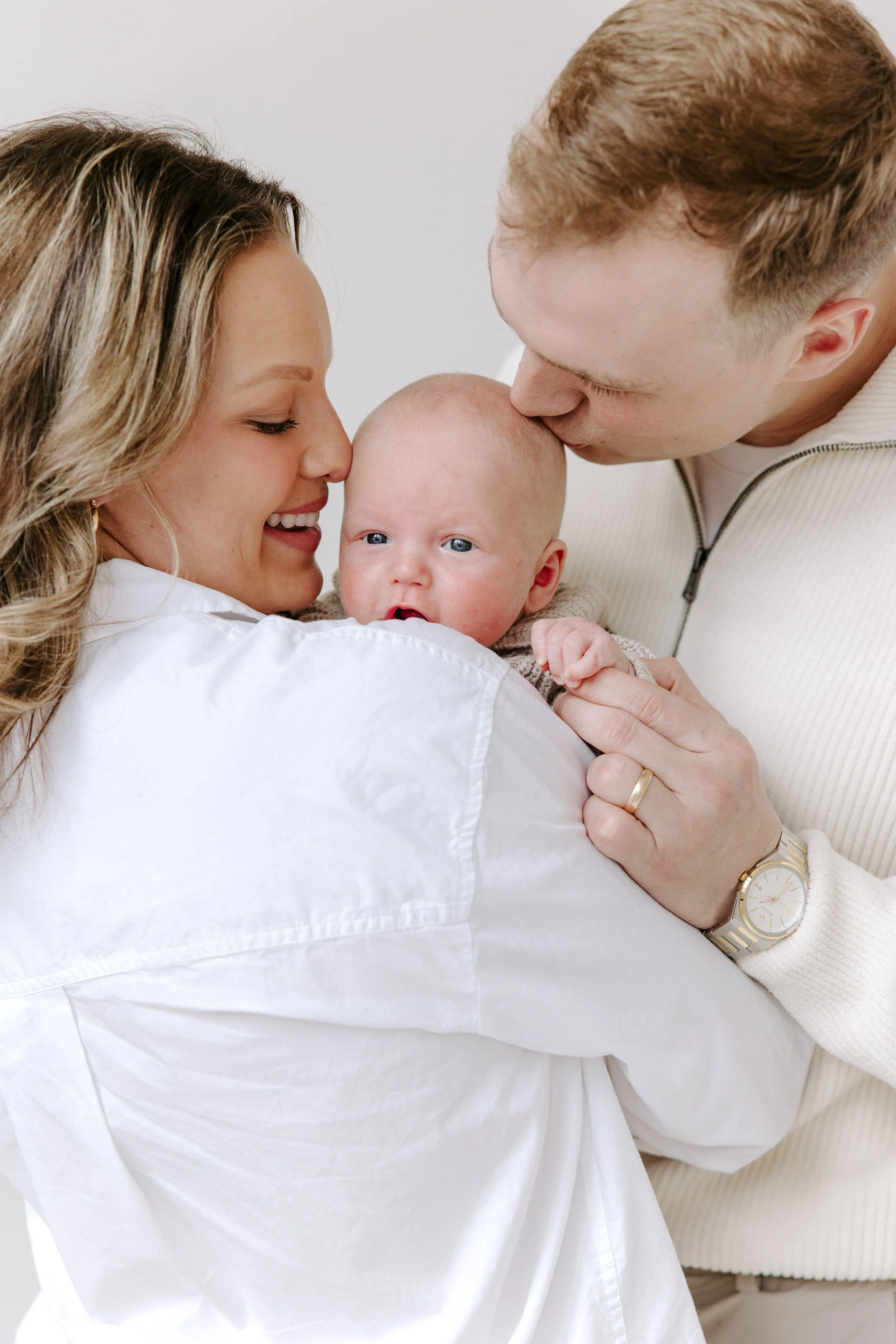 A woman and a man hold a baby close, with the man kissing the baby's forehead and the woman smiling and looking at the baby. The baby has blue eyes and is looking at the camera.