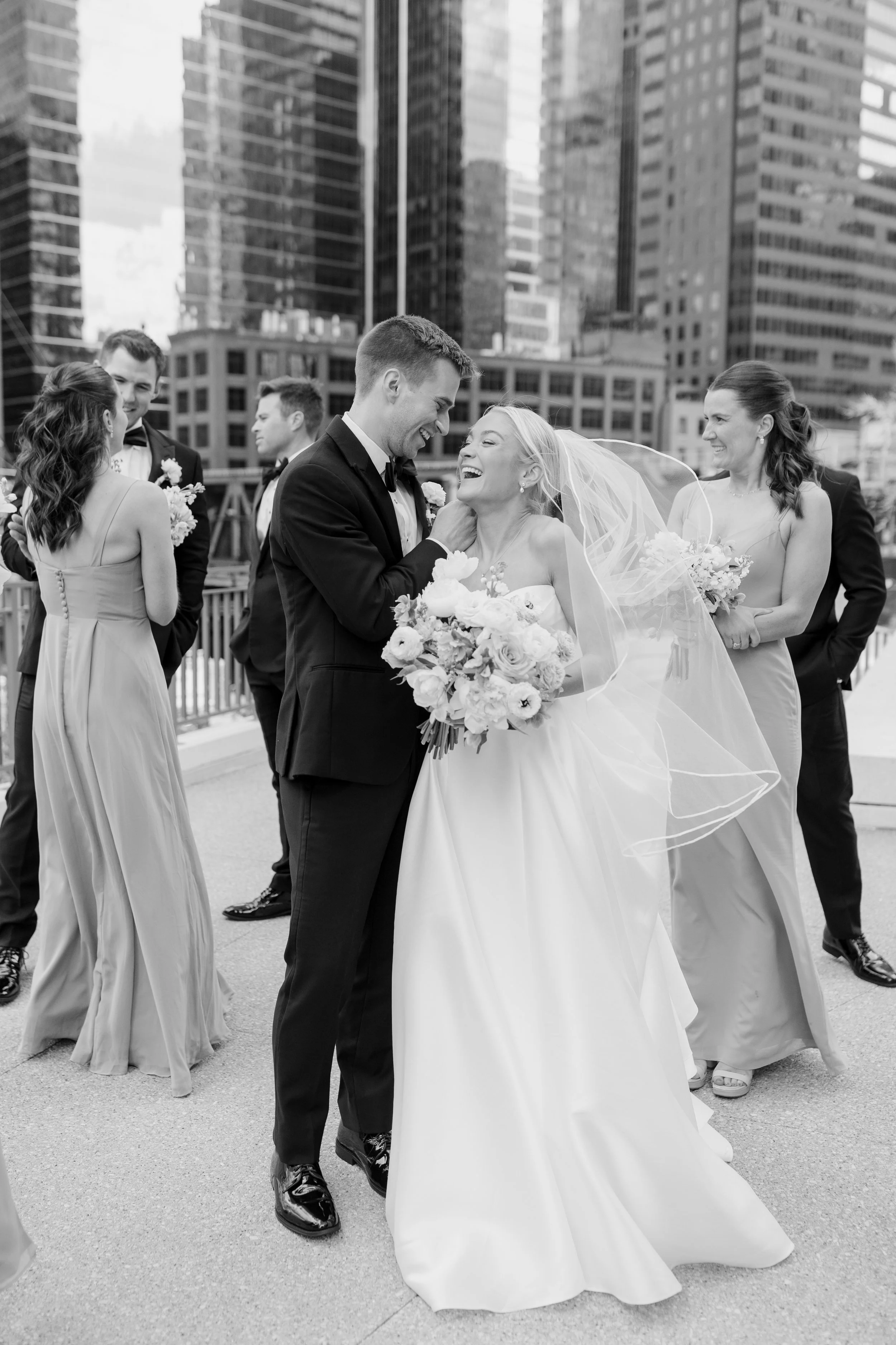 Black and white photo of a bride and groom smiling and celebrating with friends on a city rooftop. The bride, in a white wedding dress, holds a bouquet of flowers. The groom wears a black tuxedo. Four other people, three women and one man, are in the