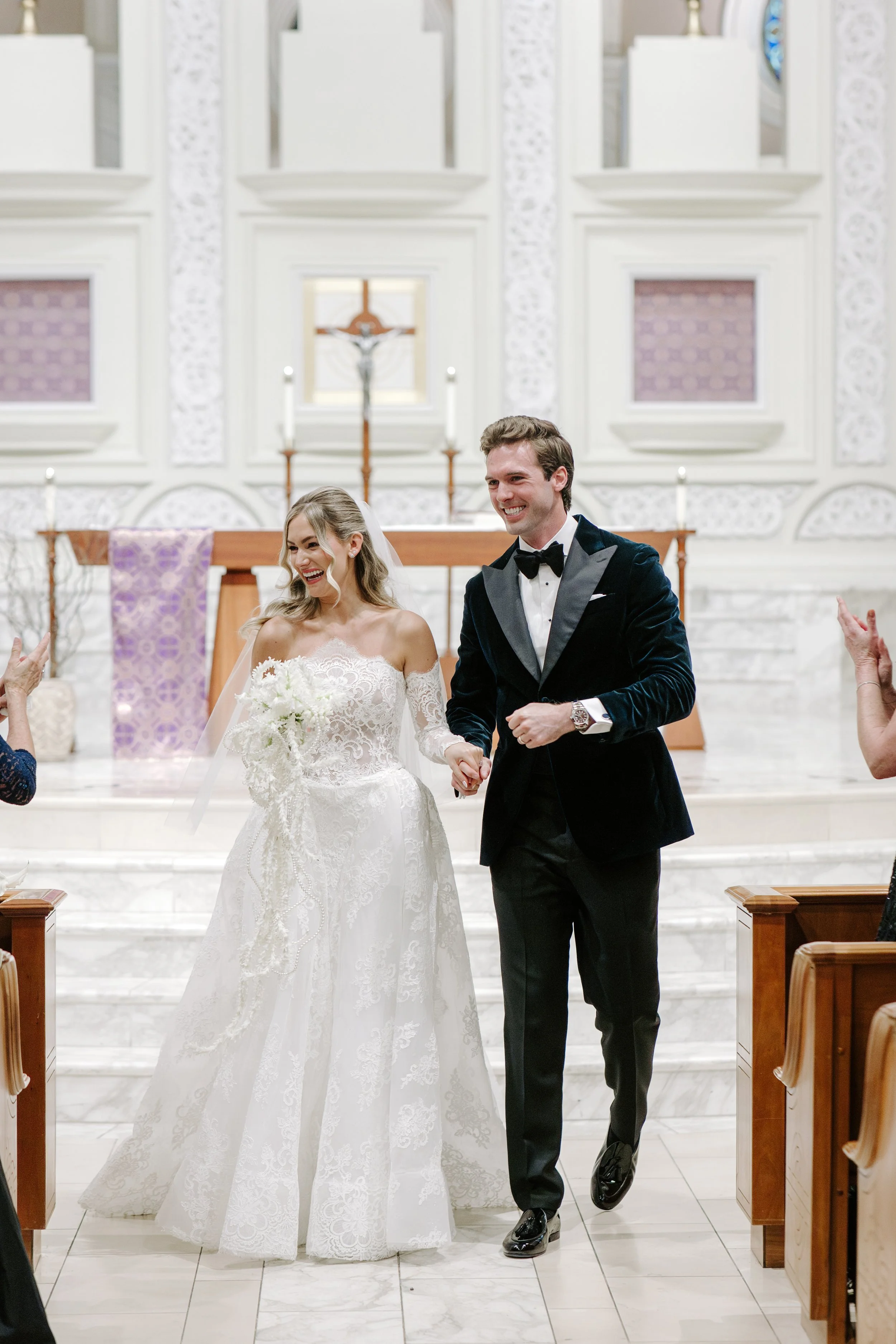 A happy bride in a white lace wedding dress holding a bouquet, and a groom in a black tuxedo with a bow tie, smiling as they walk down the aisle inside a church with white ornate walls and a crucifix in the background.