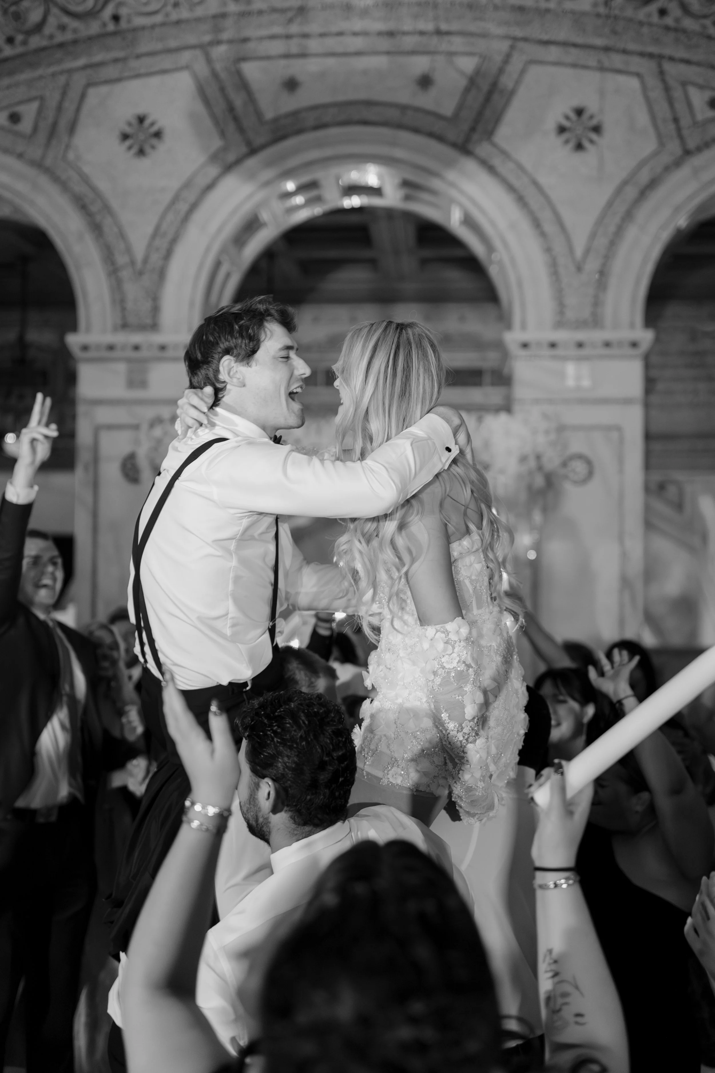 A couple dances closely during a celebration with an ornate ceiling, surrounded by guests. The woman wears a floral lace dress and the man a formal shirt with suspenders. People around them are raising hands and cheering.