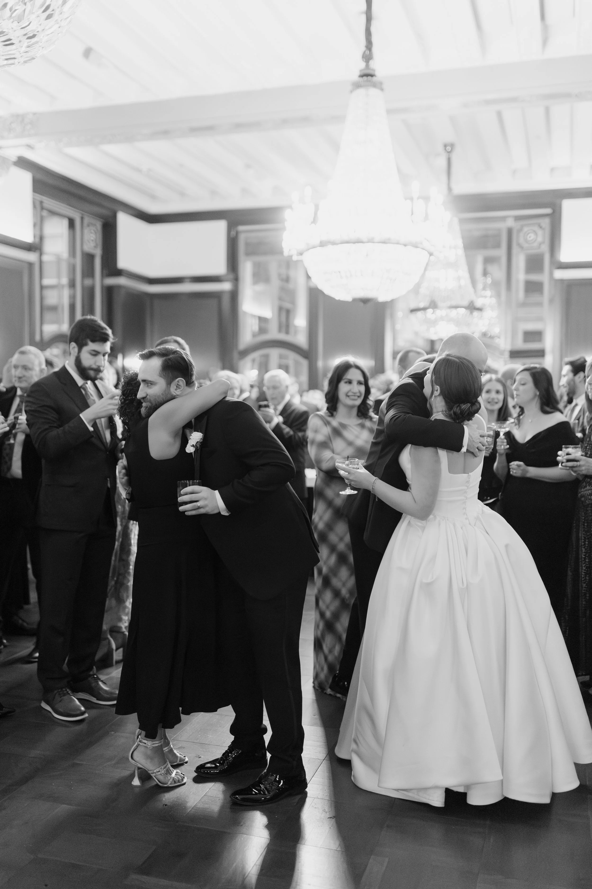 Black and white photograph of a wedding reception with people dancing, including a bride and groom embracing, surrounded by guests in formal attire in an elegant room with chandeliers.