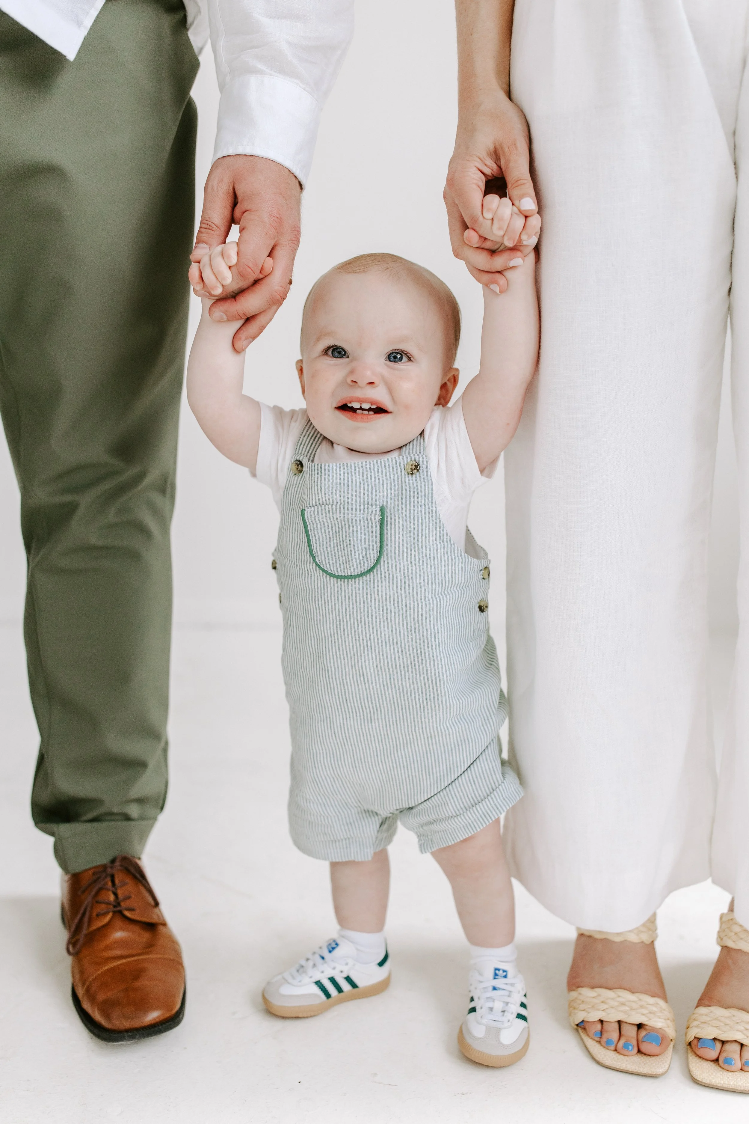 Close-up of a little boy in striped overalls holding hands with two adults, showing only their legs and hands. The adults are wearing different types of shoes and clothing.