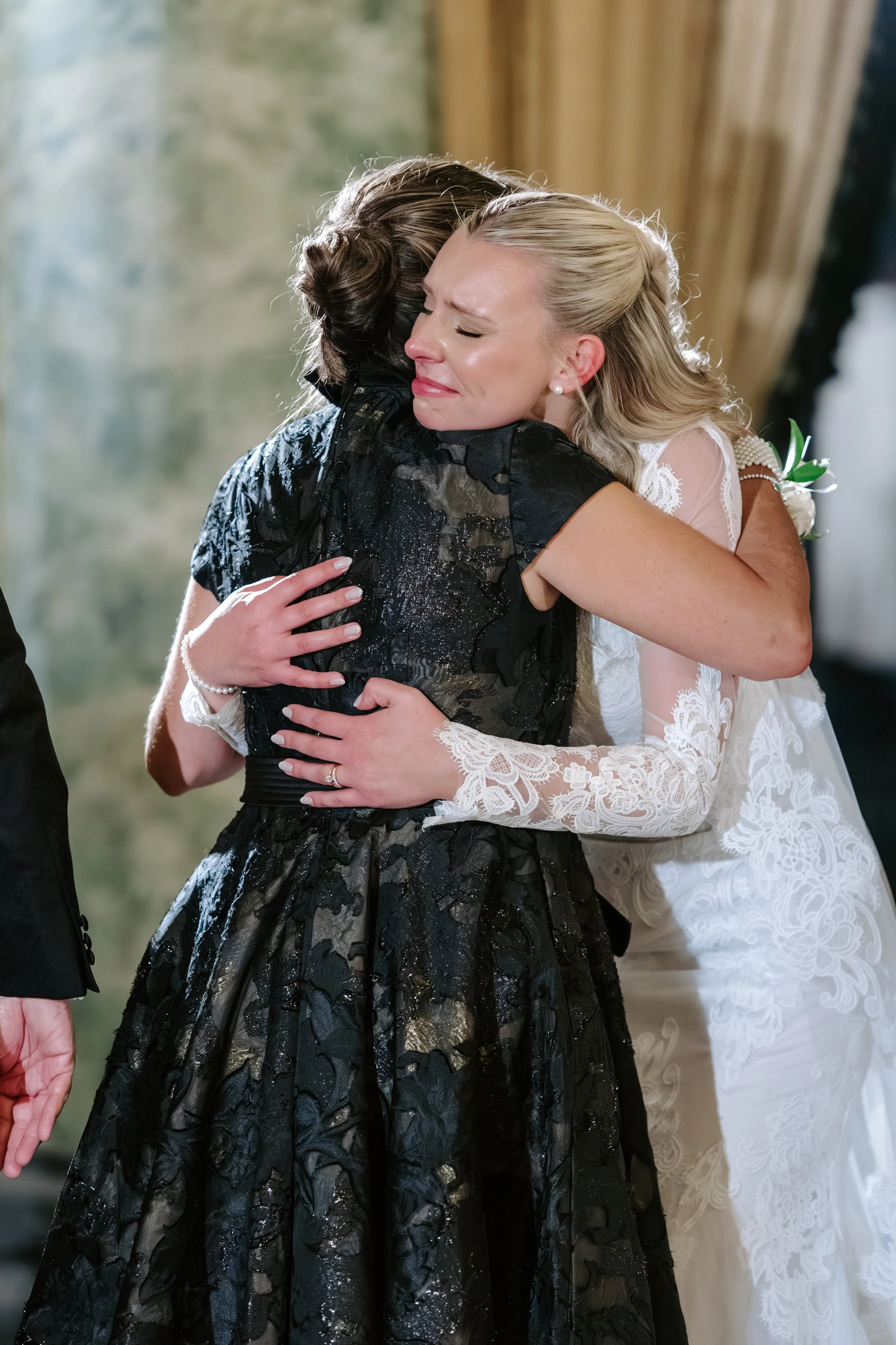 Two women embrace each other tightly, one in a black lace dress and the other in a white lace dress, during a heartfelt moment.