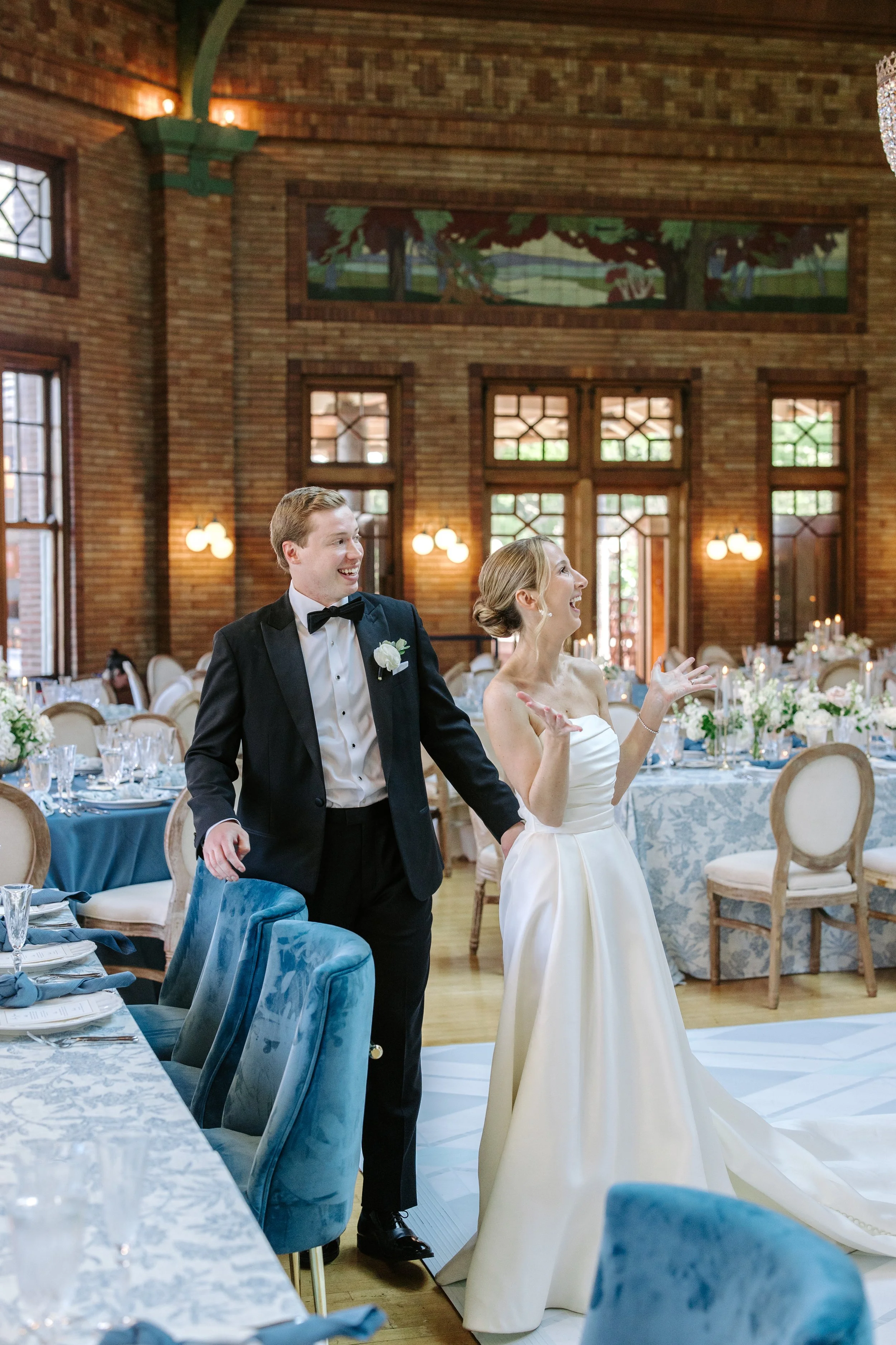 A bride and groom are dancing and smiling at their wedding reception inside a decorated venue with wooden brick walls, large windows, and chandeliers. The bride is wearing a white strapless wedding gown, and the groom is in a black tuxedo with a bow 