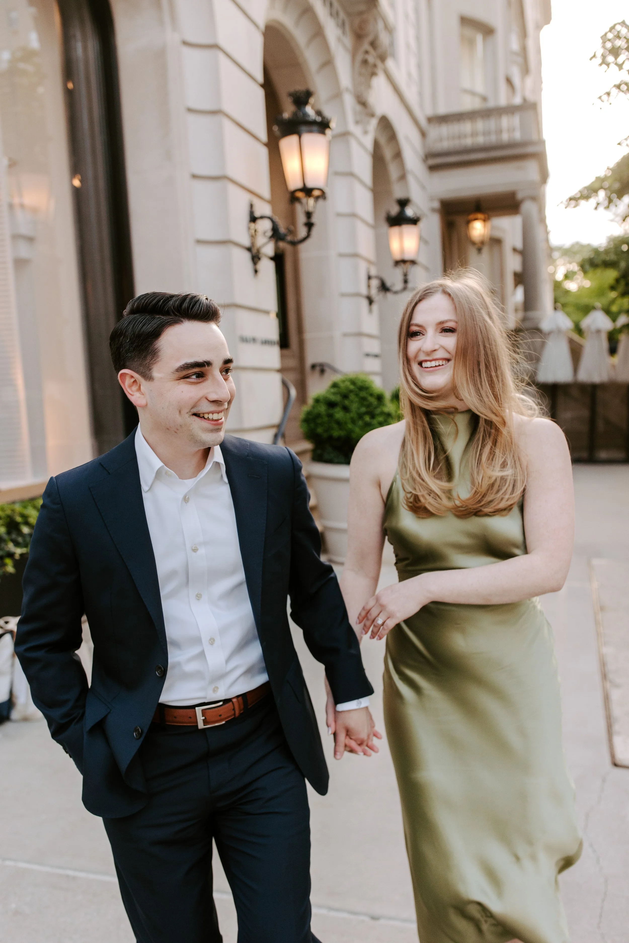A smiling couple walking hand-in-hand outside on a city street during sunset. The man is wearing a navy suit and white shirt, and the woman is in a green satin dress. Elegant city buildings with ornate lamps are in the background.