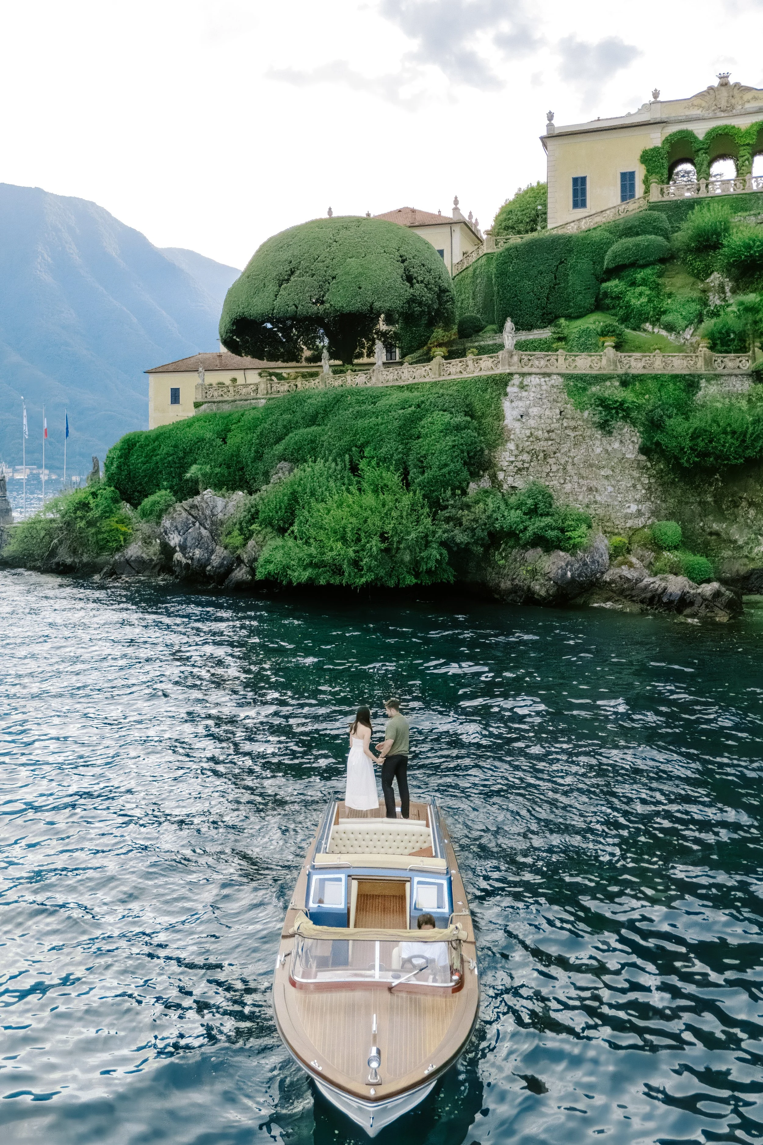 A couple holding hands stands on a small boat on a lake with a grand villa and lush terrain in the background.