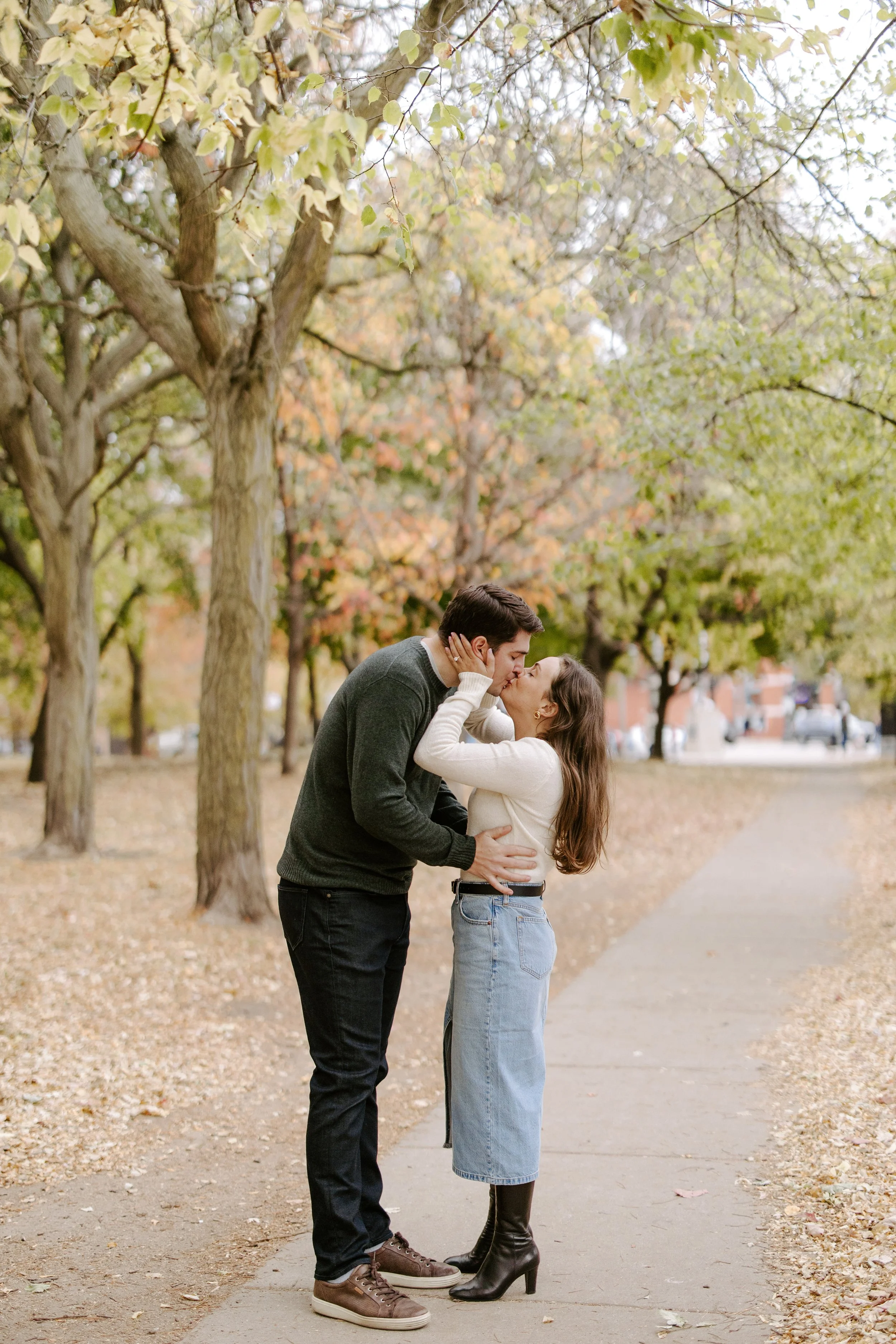 A young couple sharing a kiss in a park with trees showing fall foliage. The woman has long brown hair and is wearing a beige sweater, jeans, and black boots. The man has dark hair and is dressed in a dark sweater, black pants, and sneakers.
