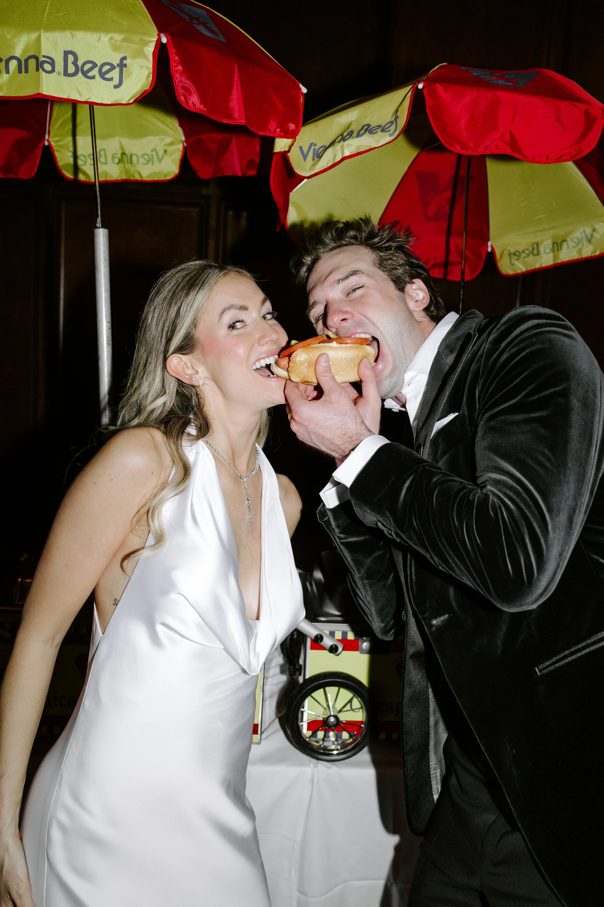 A man and a woman are sharing a large hot dog, with the man wearing a black tuxedo and the woman wearing a white dress, under red and yellow umbrellas, in an indoor setting.